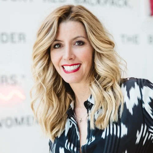 A woman with long, wavy blonde hair smiling at the camera, wearing a black and white patterned blouse, with a blurred background.