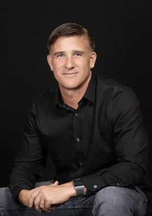 Portrait of a young man with short brown hair, wearing a black button-up shirt, sitting against a dark background.