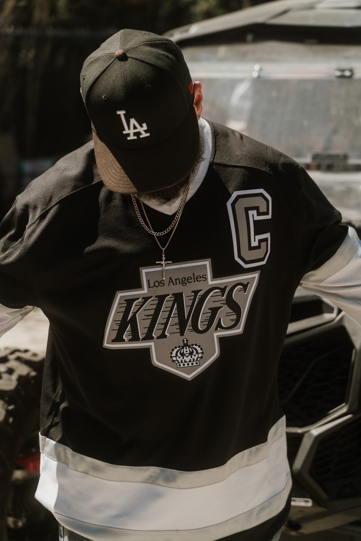Person wearing a Los Angeles Kings hockey jersey, black cap with 'LA' logo, and layered chains, standing outdoors near a vehicle.