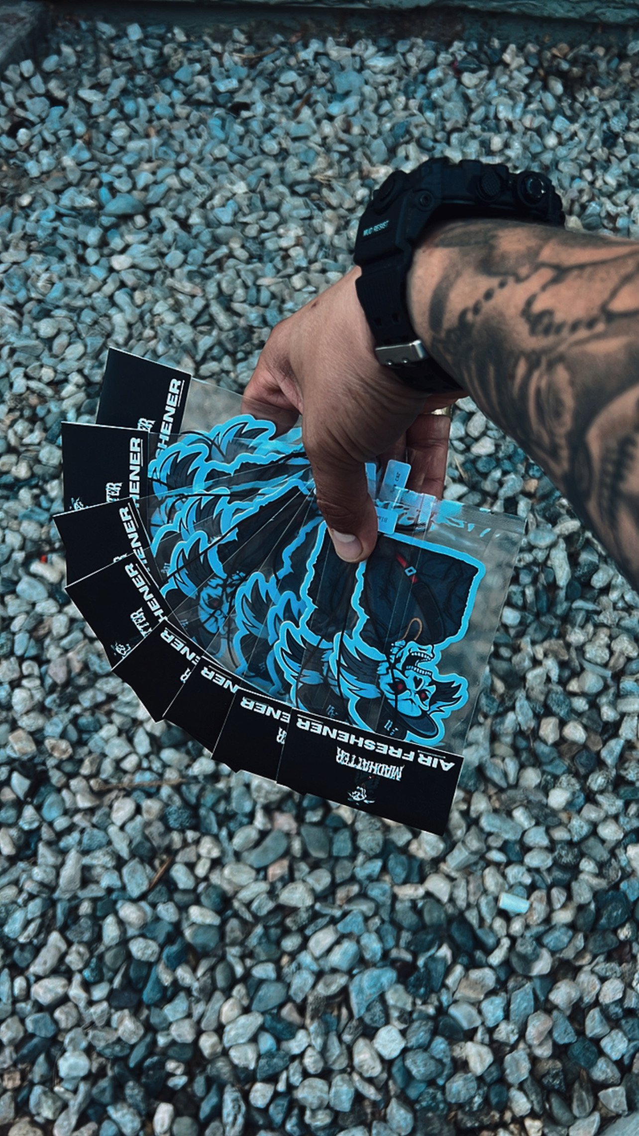 A hand holding multiple stickers featuring a blue and black wolf design, with the words "Air Feshener" on a black border around the stickers, against a gravel surface background.