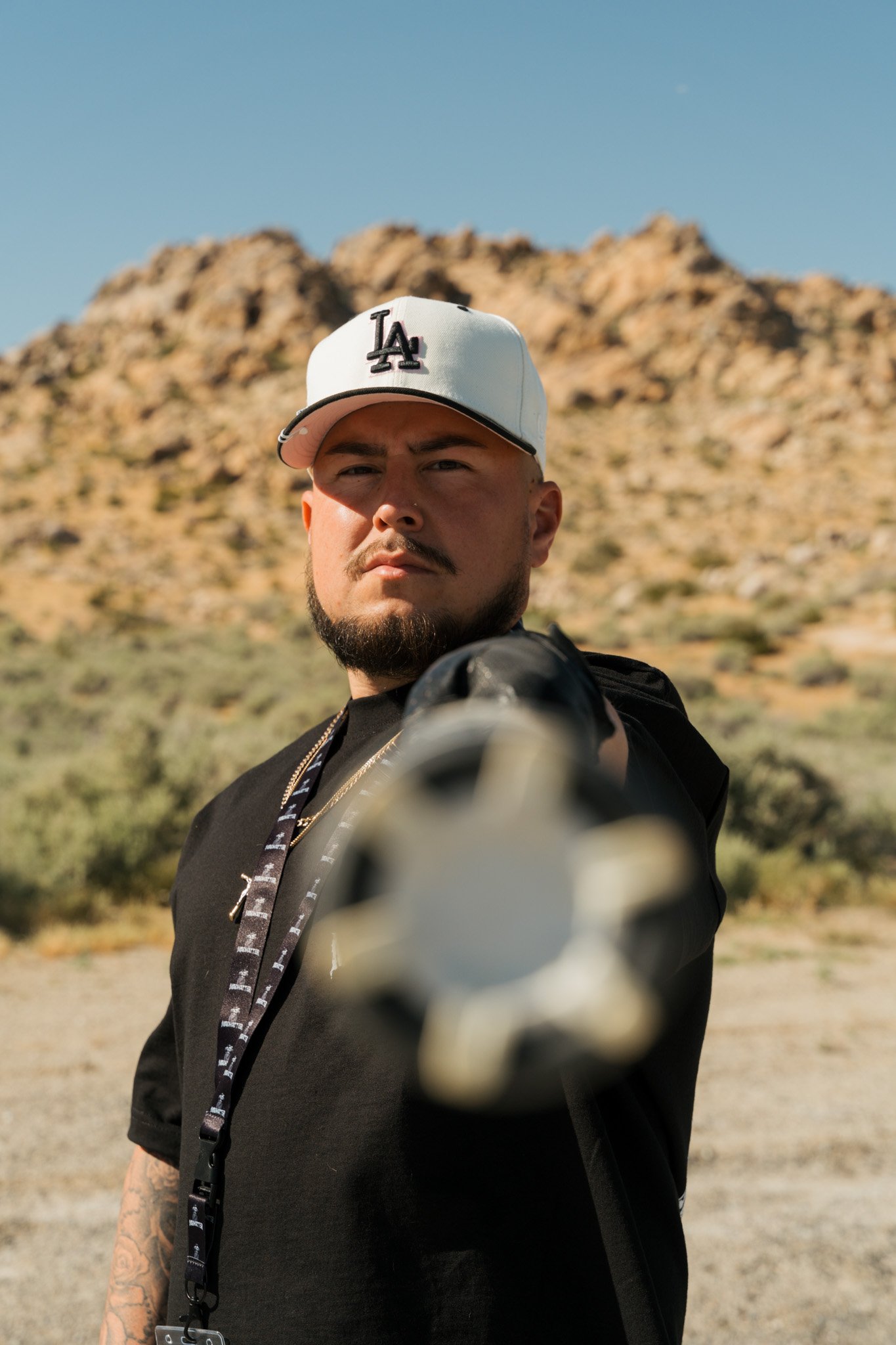 A man with a beard and mustache wearing a white Los Angeles Dodgers cap, black shirt, and a necklace points a gun directly at the camera with a desert landscape and mountains in the background.
