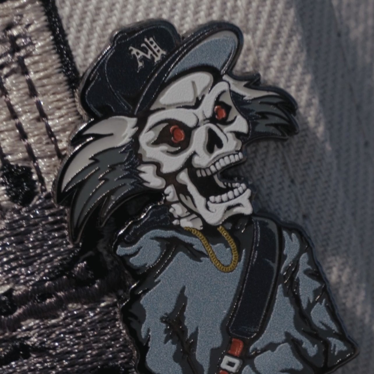 A patch or pin featuring a skeleton with a skull face, red eyes, wearing a gray hoodie, a black baseball cap, and a gold chain, on a fabric background.