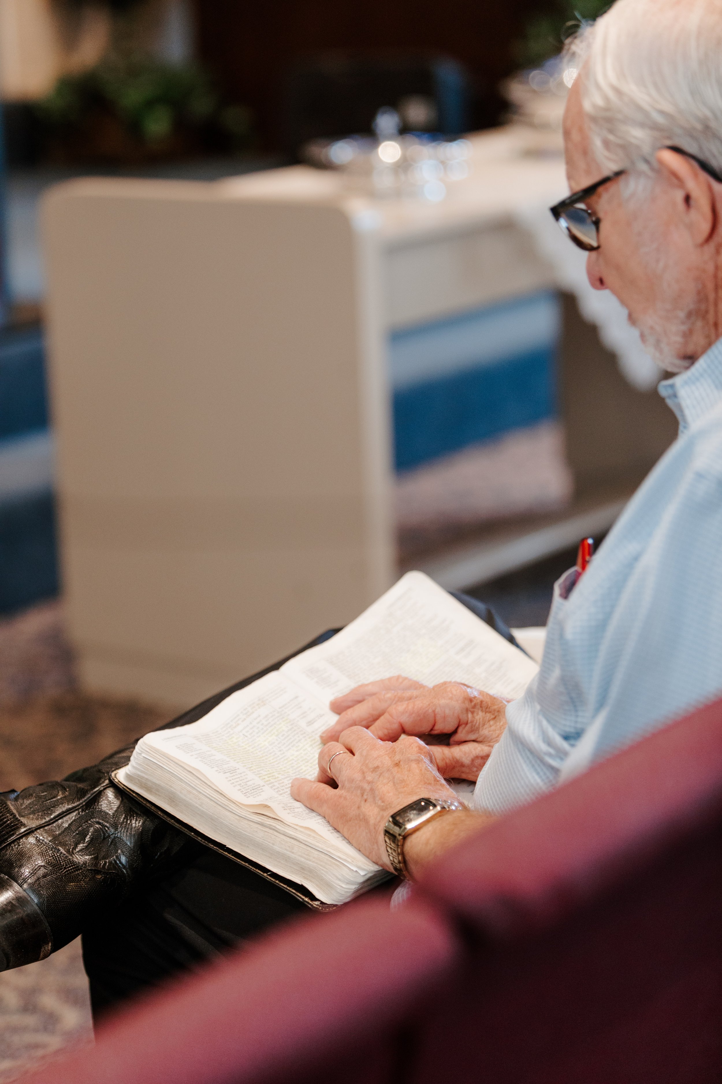 An elderly man with glasses and a white beard reading a large open Bible, sitting on a red chair.
