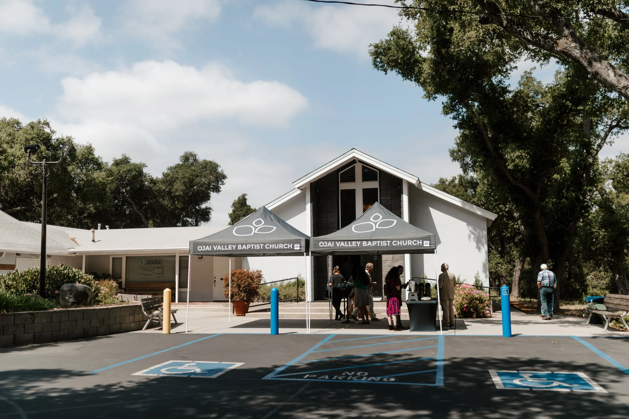 People standing outside of Ojai Valley Baptist Church under two tents with the church building in the background. There are handicap parking spaces in the foreground, some benches, and trees surrounding the area.