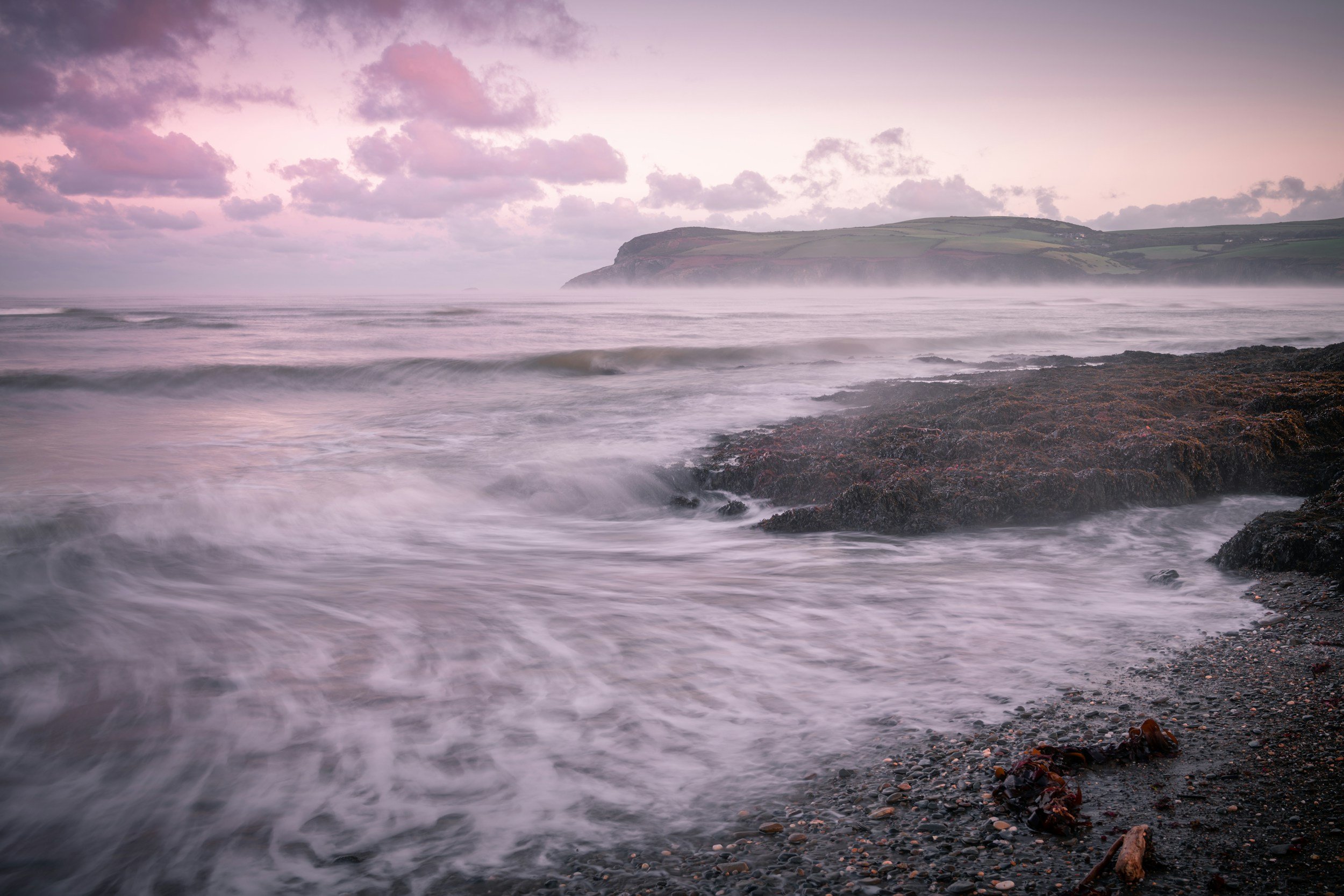 Soft lavender light over a misty ocean and rocky coast at sunrise.