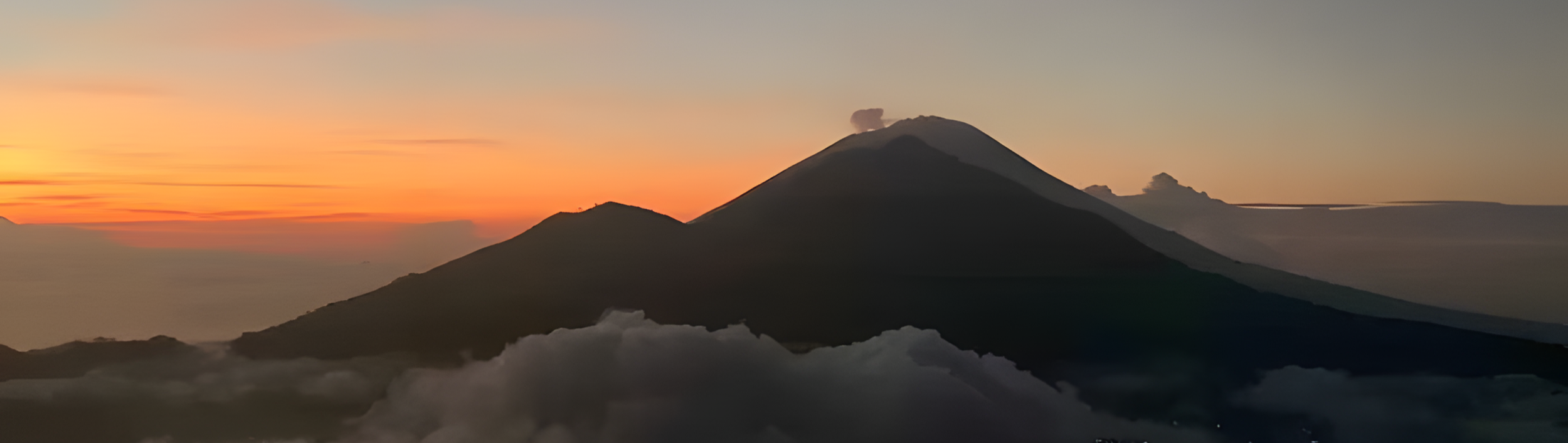 A scenic view of a volcanic landscape at sunset with a prominent volcano emitting a small puff of smoke, colorful sky with orange and pink hues, and clouds partially covering the base of the volcano.
