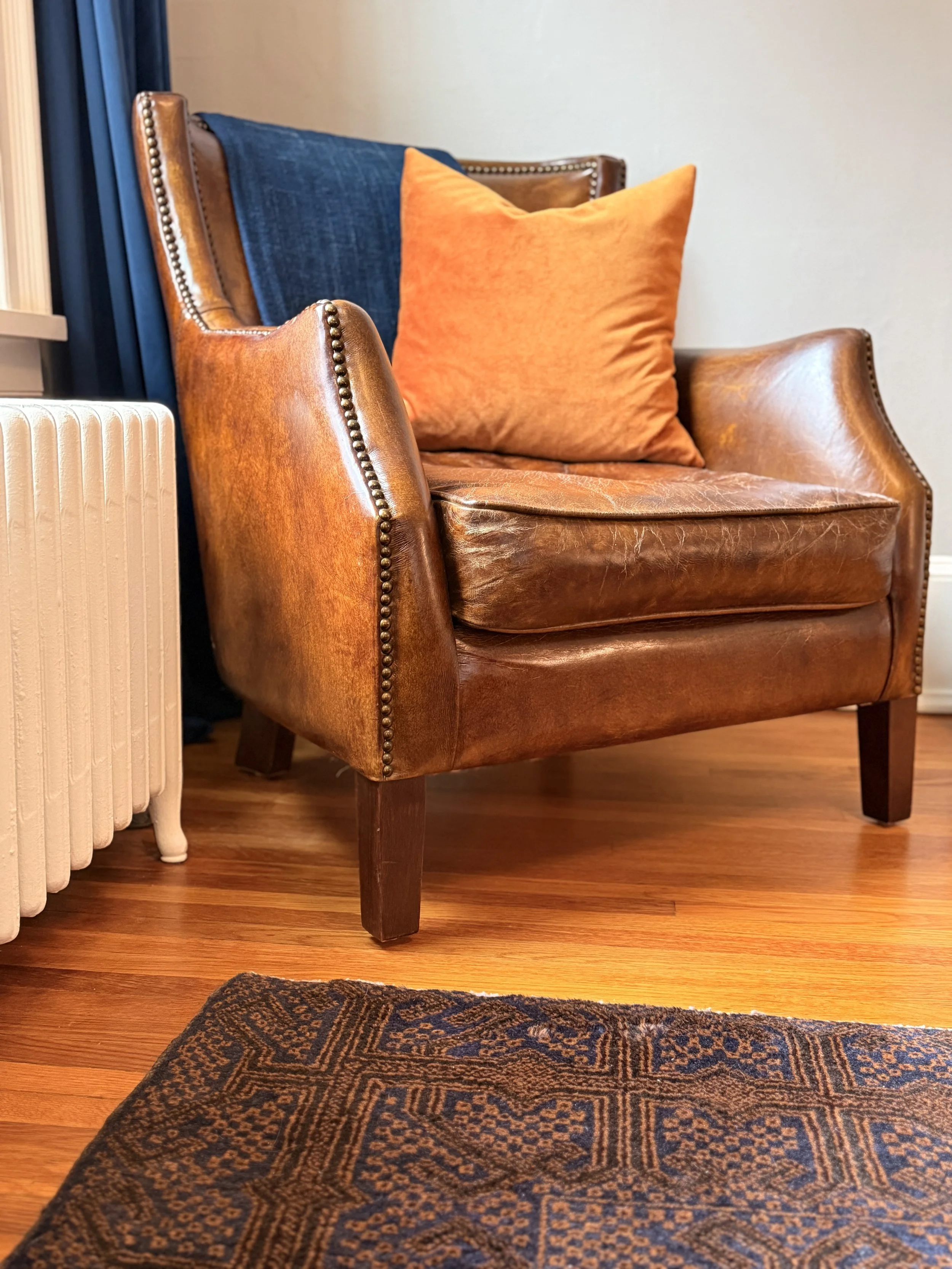 A vintage brown leather armchair with a large orange pillow, a blue cushion on the backrest, and wooden legs, situated next to a white radiator and on a wooden floor, with a patterned rug partially visible in the foreground.