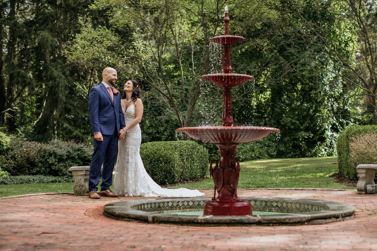 A bride and groom dressed in wedding attire standing hand in hand outdoors near a red fountain, with green trees and bushes in the background.