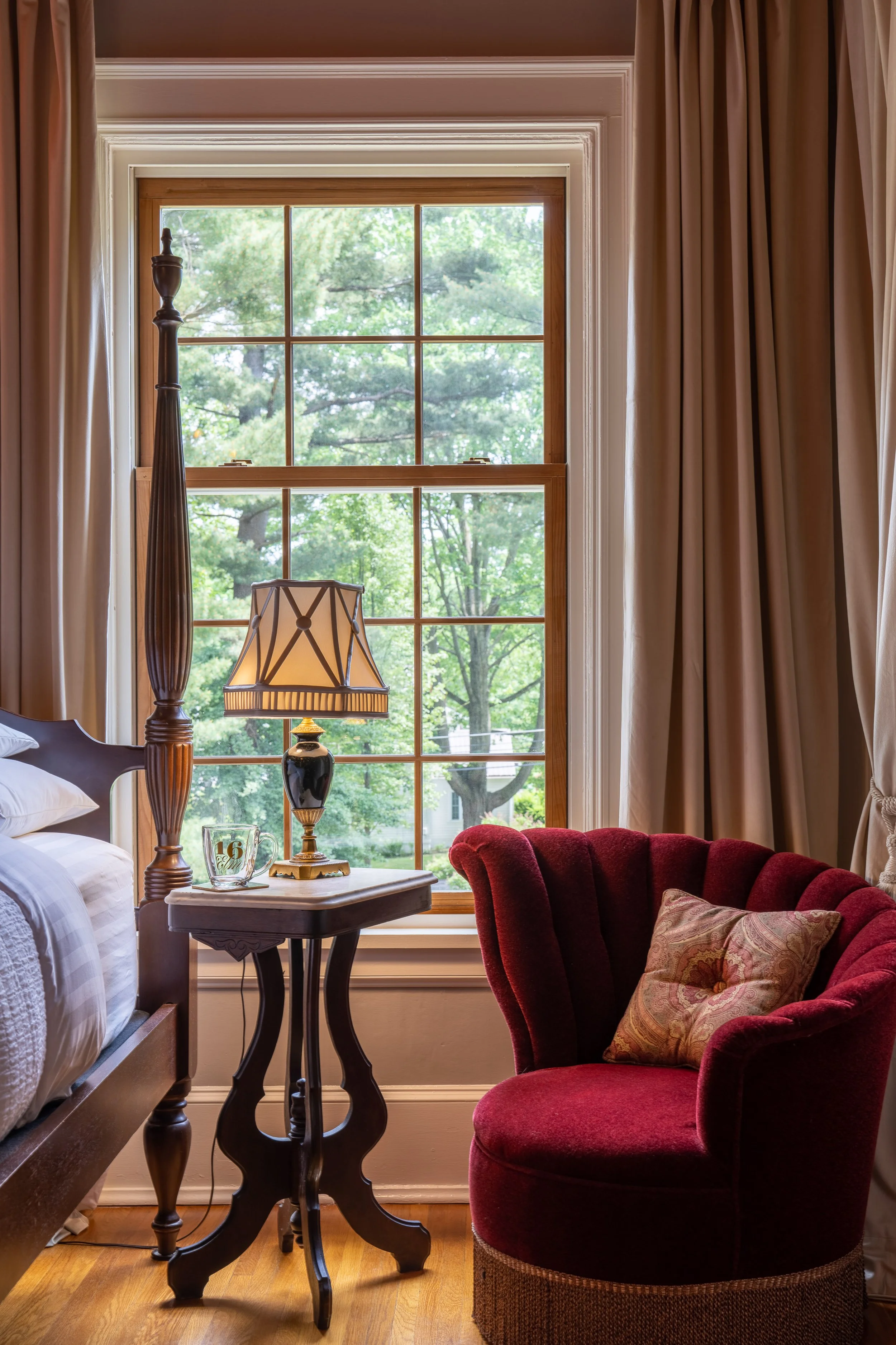 A bedroom corner with a large window, a bedside table with a lamp, a red velvet armchair with a throw pillow, and cream-colored curtains.