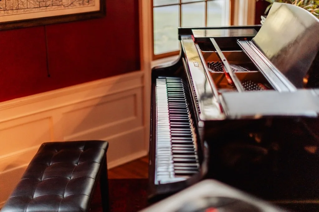 An upright black grand piano in a room with a black padded piano bench, wooden wall paneling, a window, and a framed picture on the wall.
