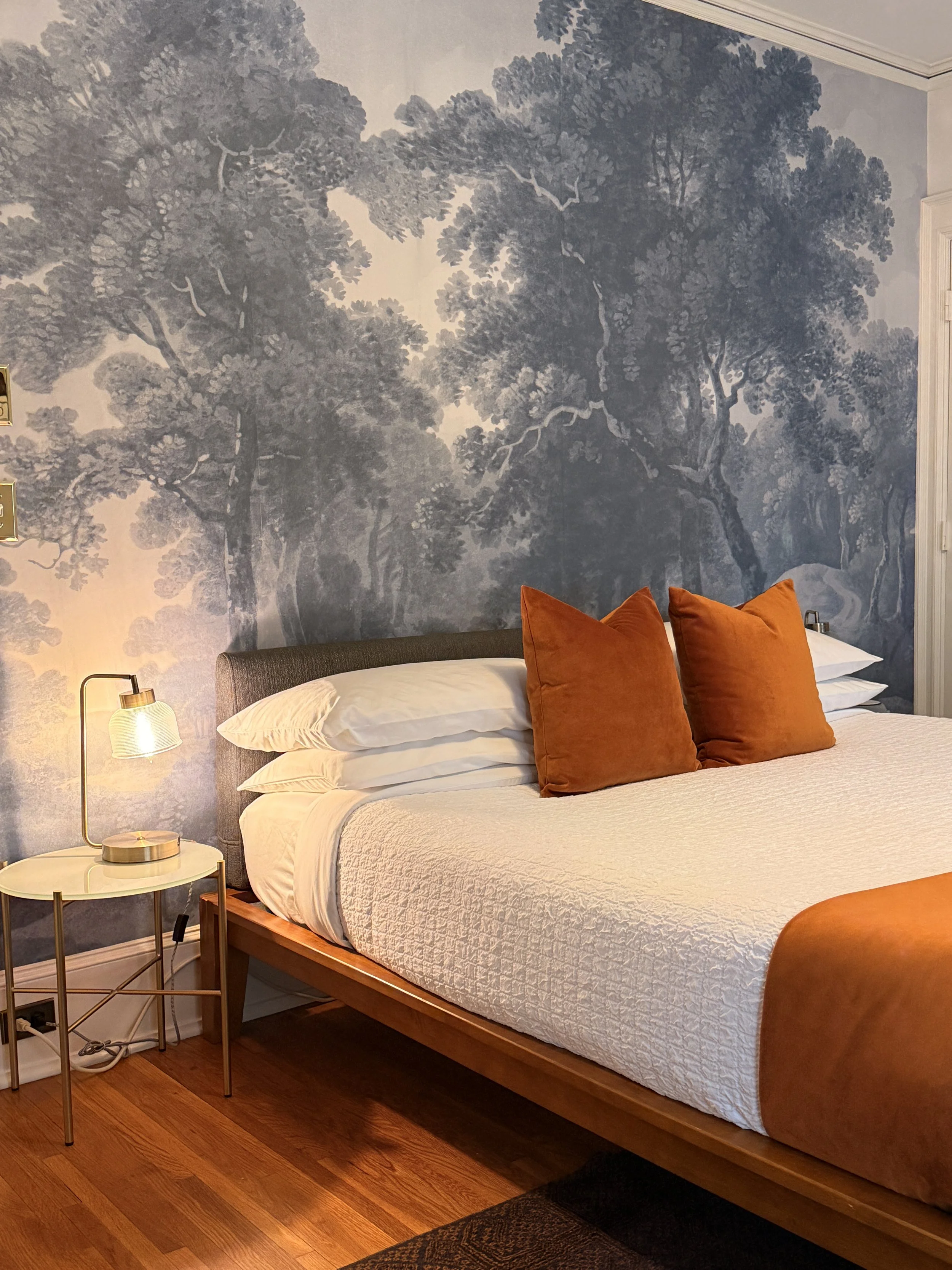 A bedroom with a headboard, white bedding, two orange pillows, a small white bedside table with a lamp, and a blue mural of trees on the wall behind the bed.