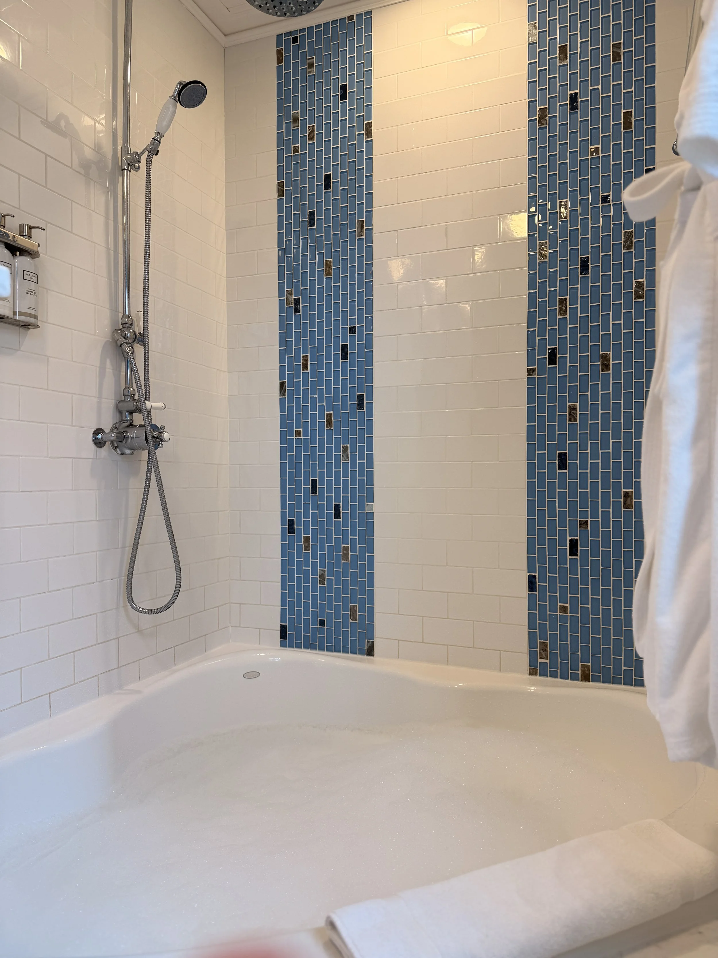 White bathtub with a gentle slope, soap suds on the surface, a white towel resting on the edge; white tiled wall with vertical blue mosaic tiles in the background; showerhead and fixtures on the left wall, a small soap dispenser on the left side.