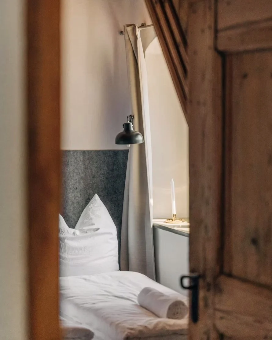 Ein gemütliches Schlafzimmer mit einem Bett, weißen Bettwäsche, einem weißen Kissen, einer schwarzen Wandlampe, weißen Vorhängen und einer Kerze auf dem Fensterbrett, sichtbar durch eine offene Holztür.