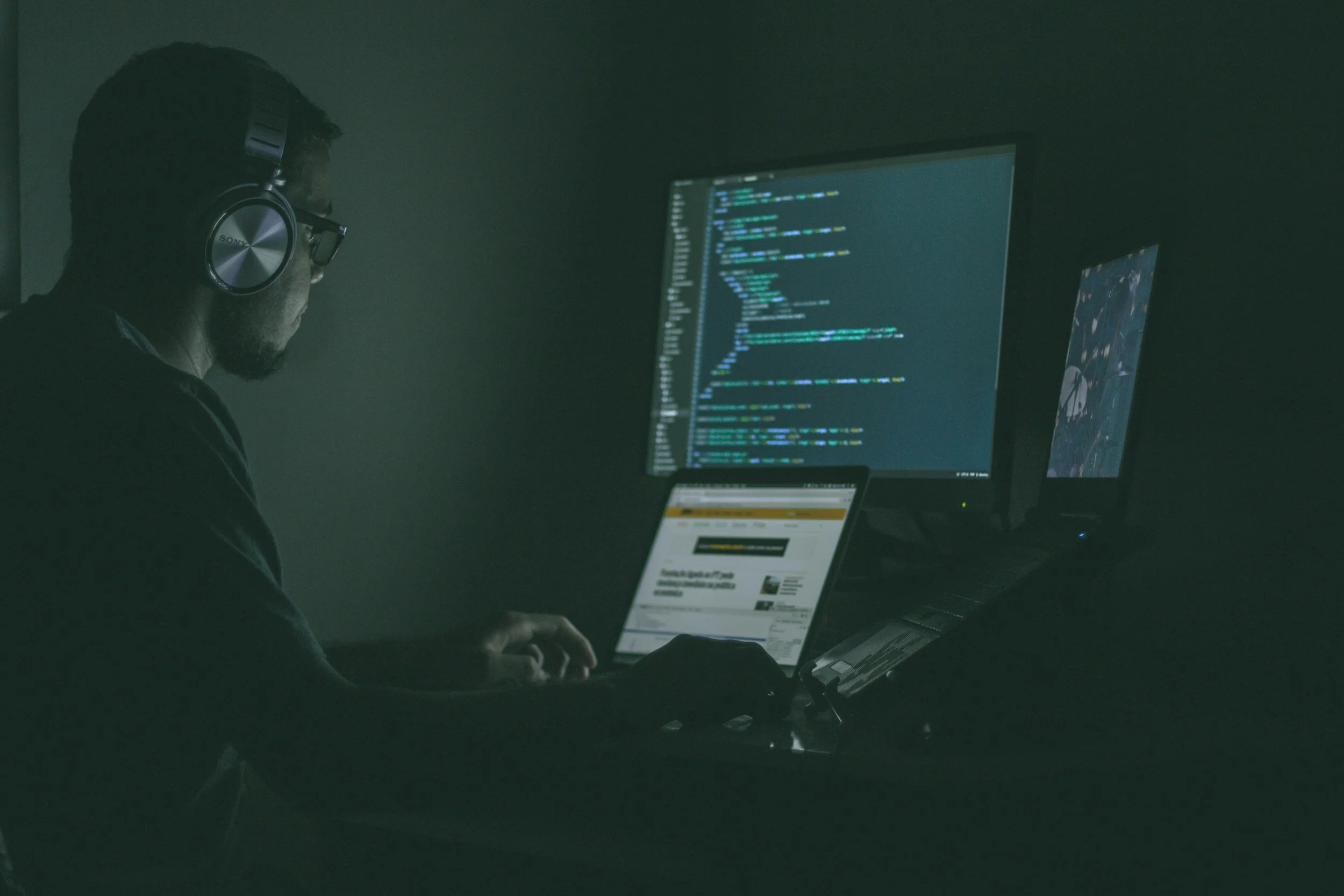 A person wearing headphones working on multiple computer screens displaying code in a dimly lit room.