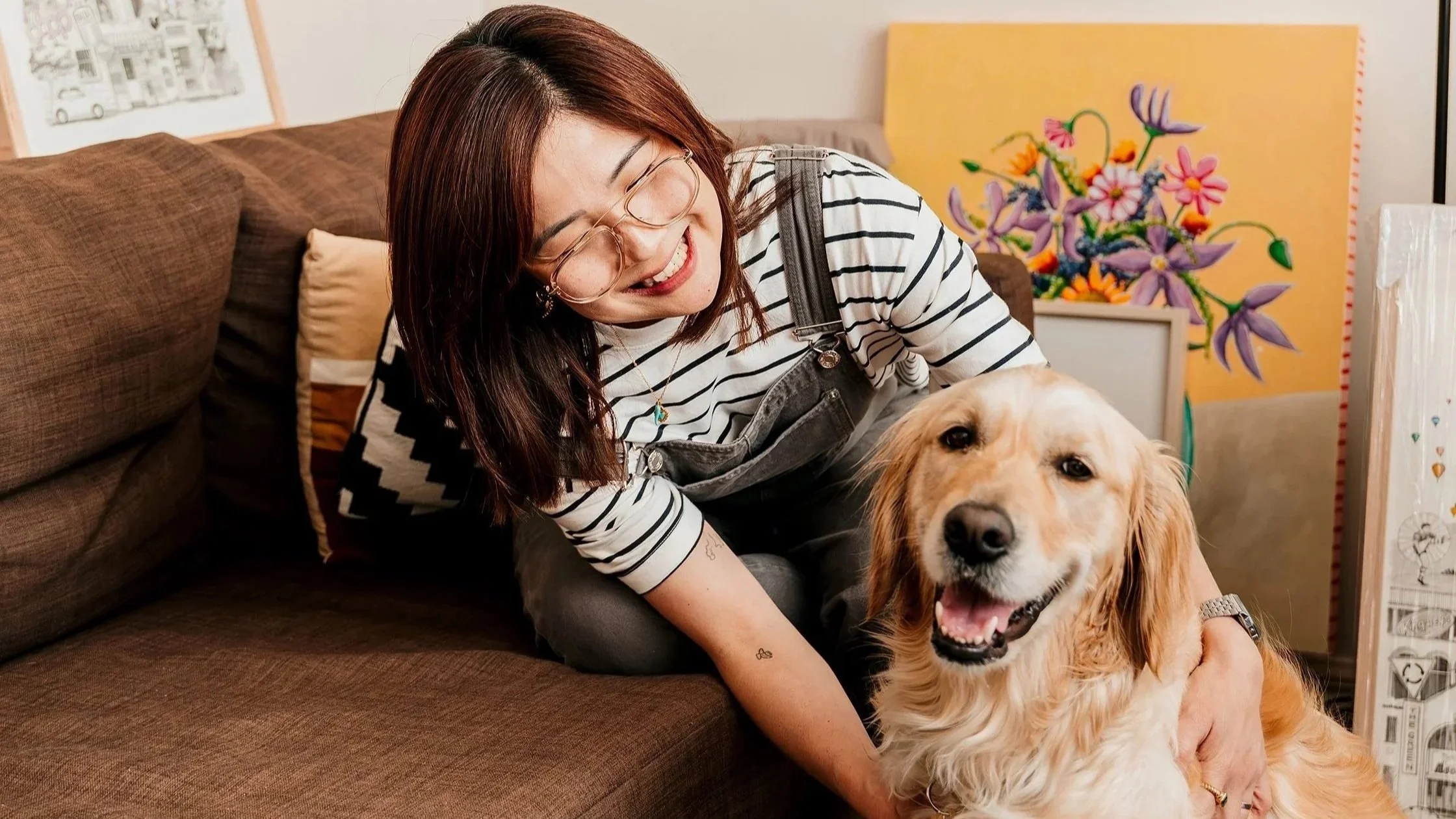 A woman with red hair, glasses, and striped shirt smiling as she bends down to hug a happy golden retriever dog on a brown couch surrounded by artworks.