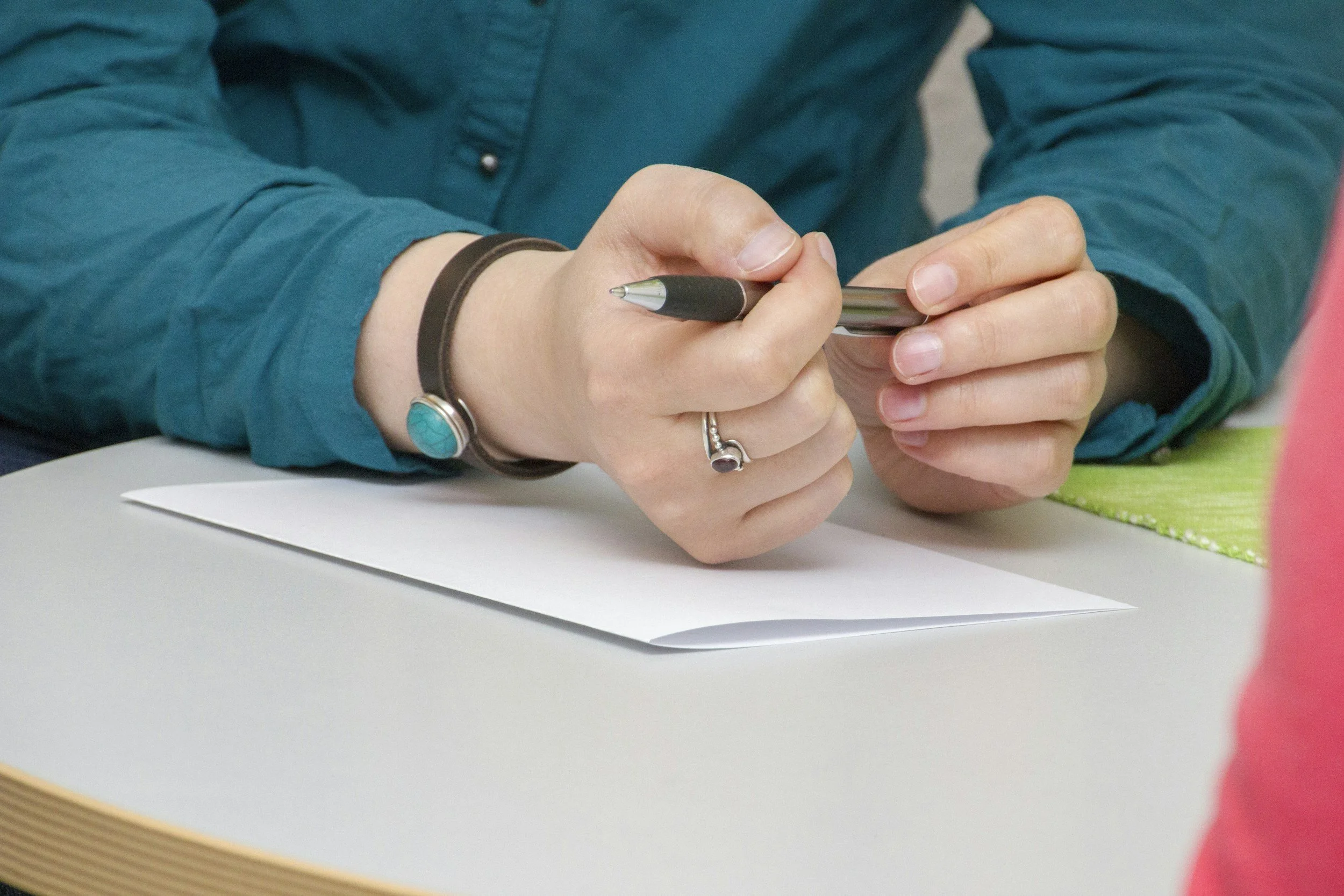 A person wearing a teal shirt is holding a black marker and writing on paper against a light-colored table.