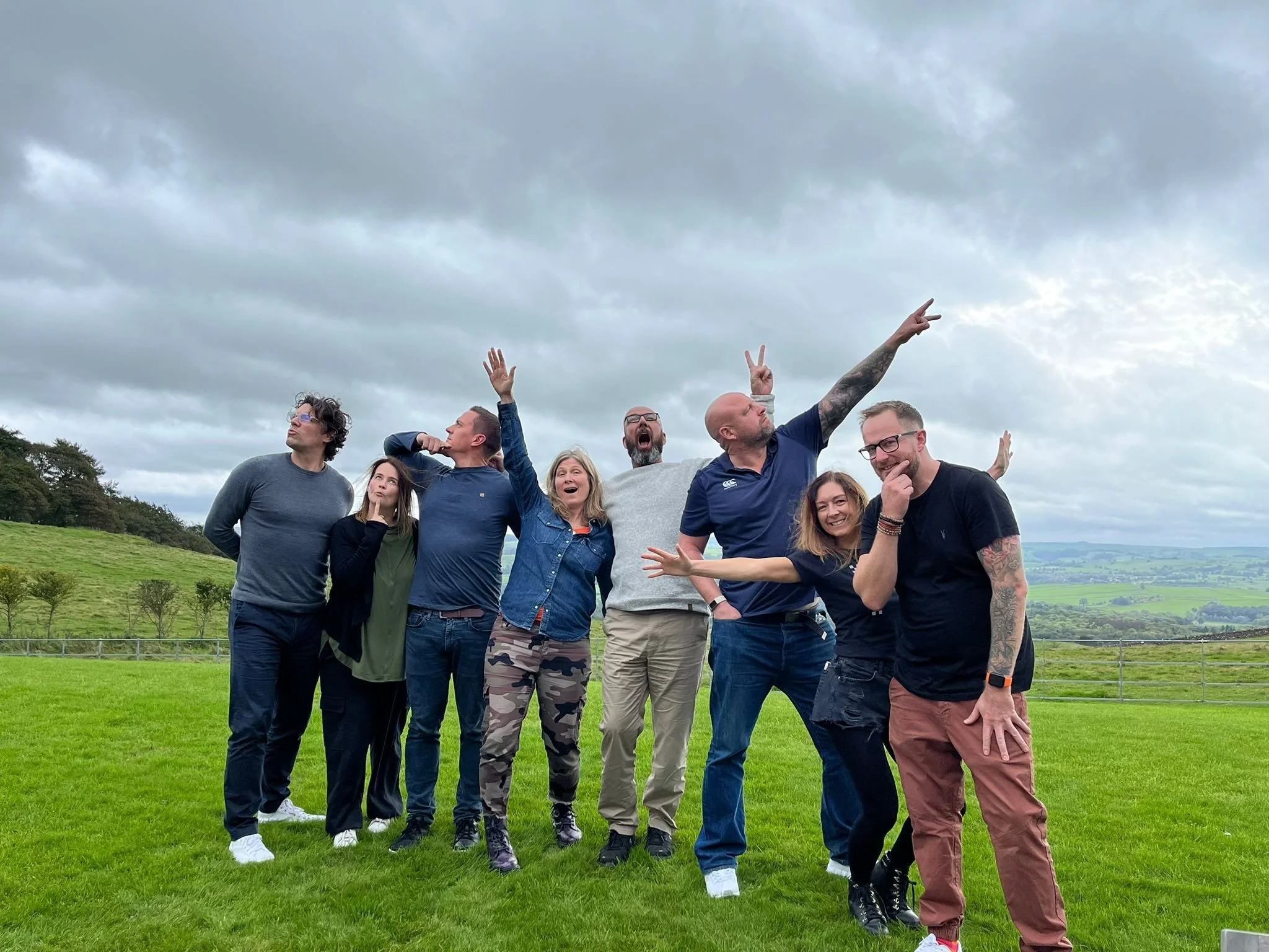 A group of people from the retreat, standing on a grassy field outdoors under a cloudy sky, posing and making gestures for the camera.