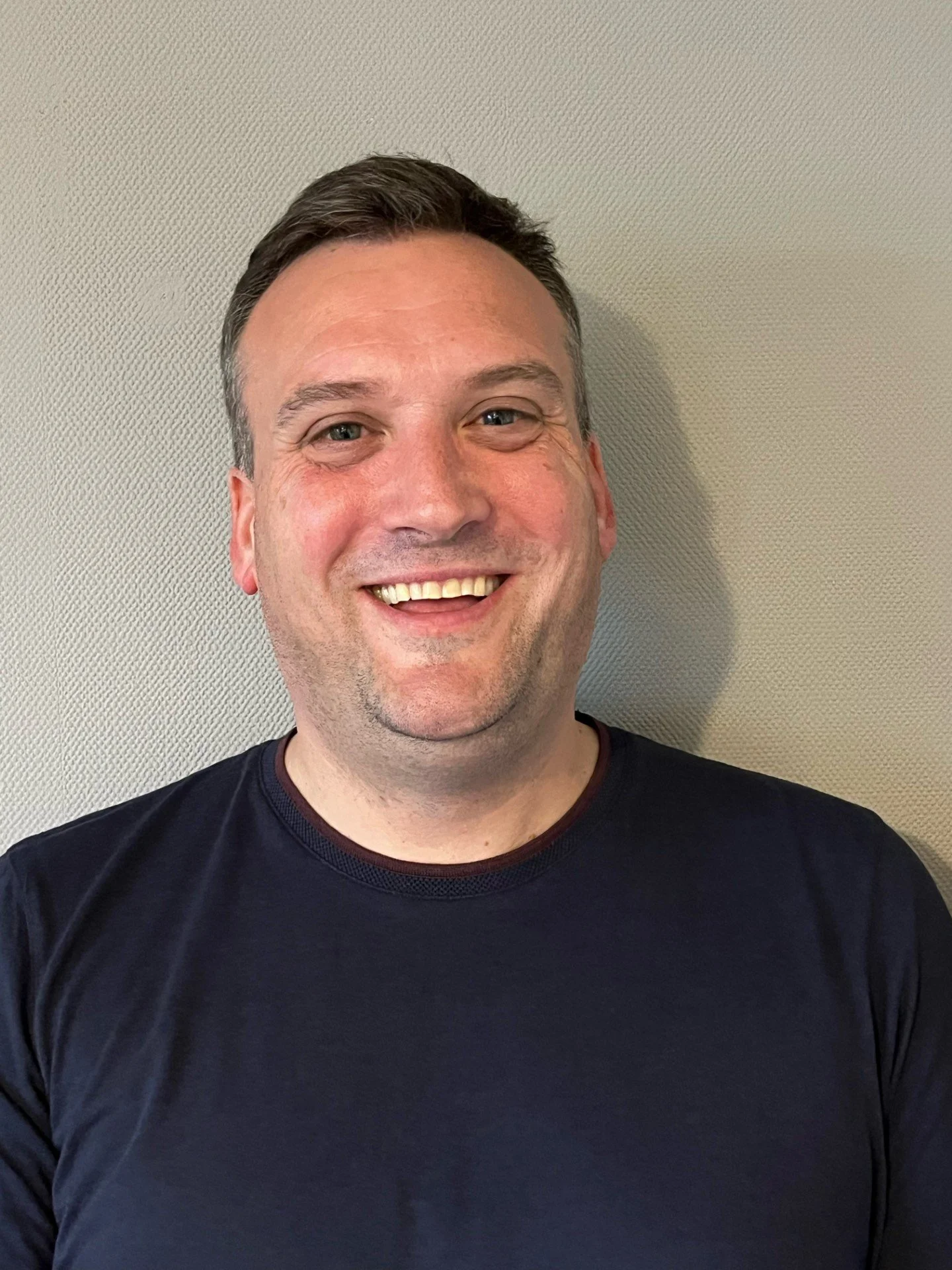 A portrait of Michael, a smiling man with short dark hair, wearing a black T-shirt, against a beige textured wall.