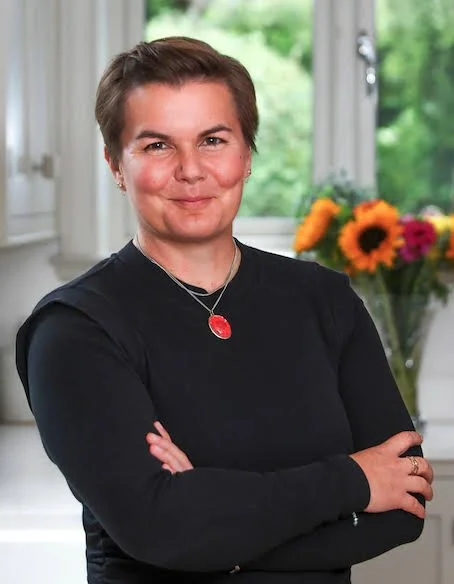 A portrait of Gemma, a woman with short hair wearing a black top and a red pendant necklace, standing with arms crossed in a bright kitchen with a window and a vase of colourful flowers in the background.