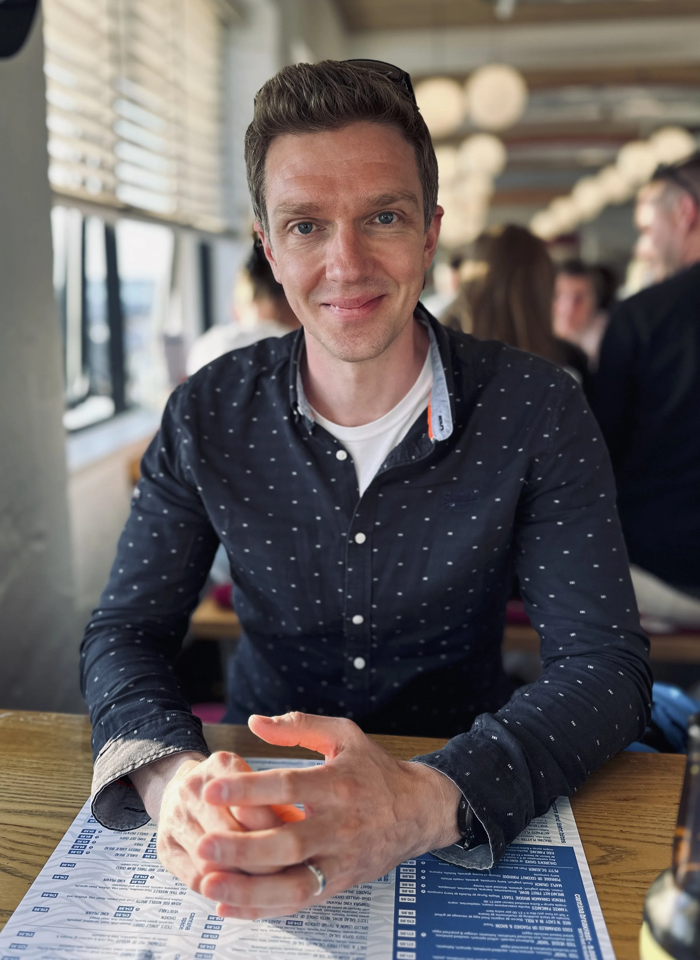 A portrait of Gareth, our designer. He has short, blonde hair and is wearing a dark shirt with small white dots. The background shows other people and a window with blinds.