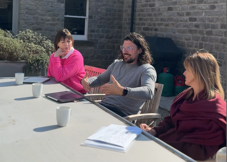 A retreat group of three people sitting at an outdoor table having a conversation, with a brick house and garden in the background.