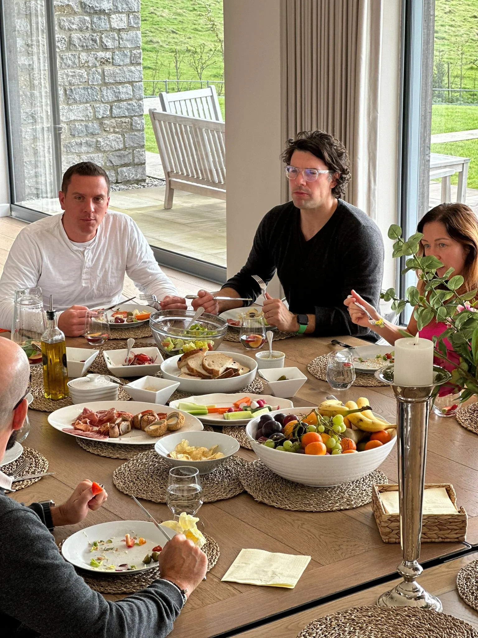 A retreat group gathering at a dining table with various foods including bread, fruits, vegetables, and salads, with three visible people eating and enjoying their meal in a bright room with large windows showing a green outdoor landscape.