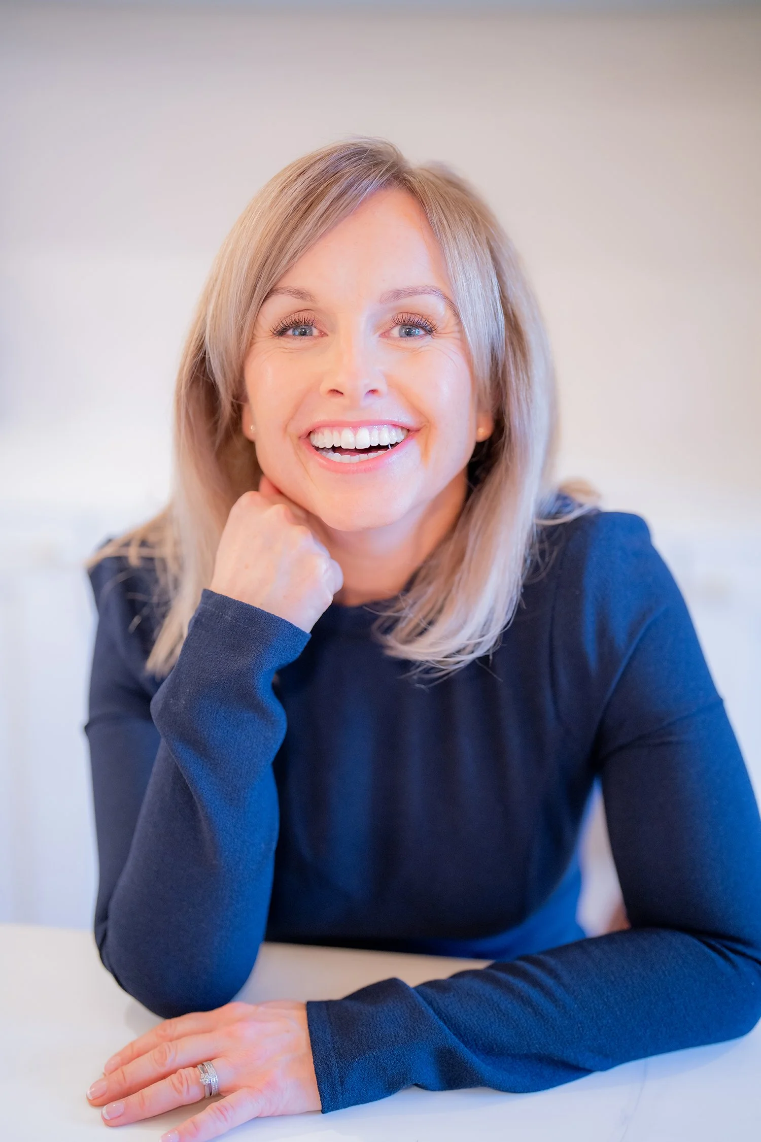 A portrait of Nicky, smiling with blonde hair, sitting at a table, resting her chin on her hand, wearing a dark long-sleeve top, with a wedding ring on her ring finger.
