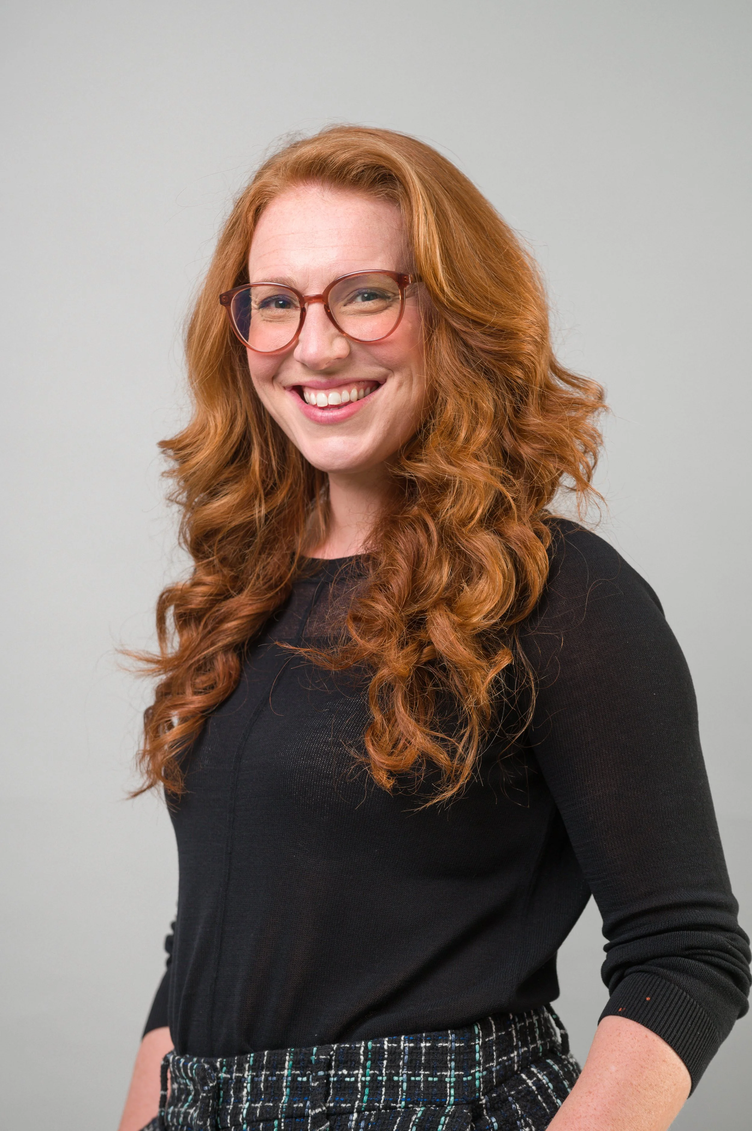 A portrait of Rebecca, a woman with long curly red hair wearing glasses, a black top, and patterned pants, smiling against a plain light grey background.