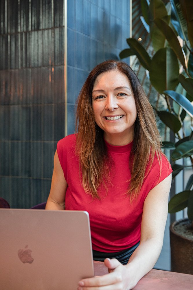 A portrait of Laura, a woman with shoulder-length brown hair smiling at the camera, sitting at a table with a pink MacBook laptop, in a modern indoor setting with large green plants and blue-tiled walls.