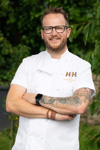 A portrait of Mike, our chef with glasses, tattoos, and a smartwatch, wearing a white chef's coat with the HH logo, standing outdoors with greenery in the background.
