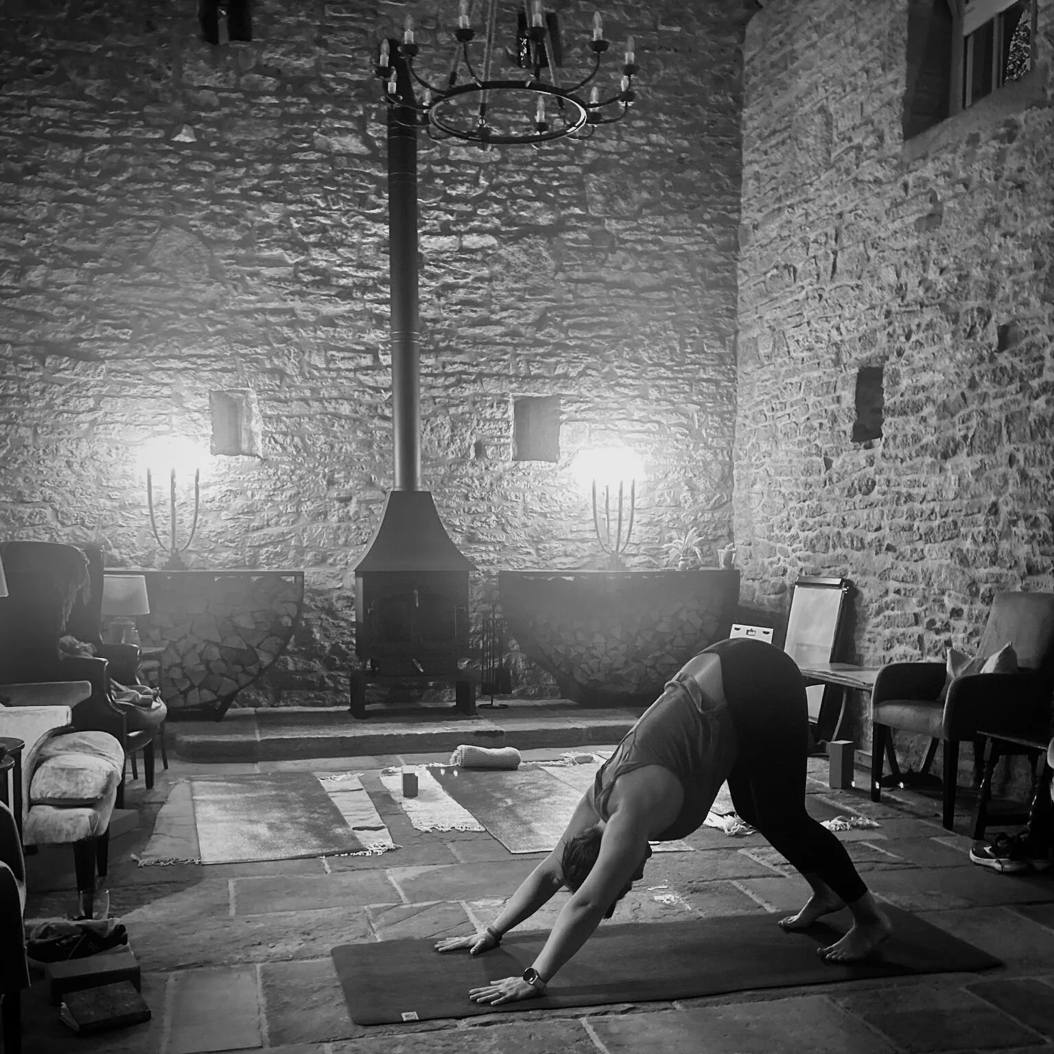 A person practising yoga in a rustic room with stone walls and a wood-burning stove, in a downward dog pose on a yoga mat during our retreats.