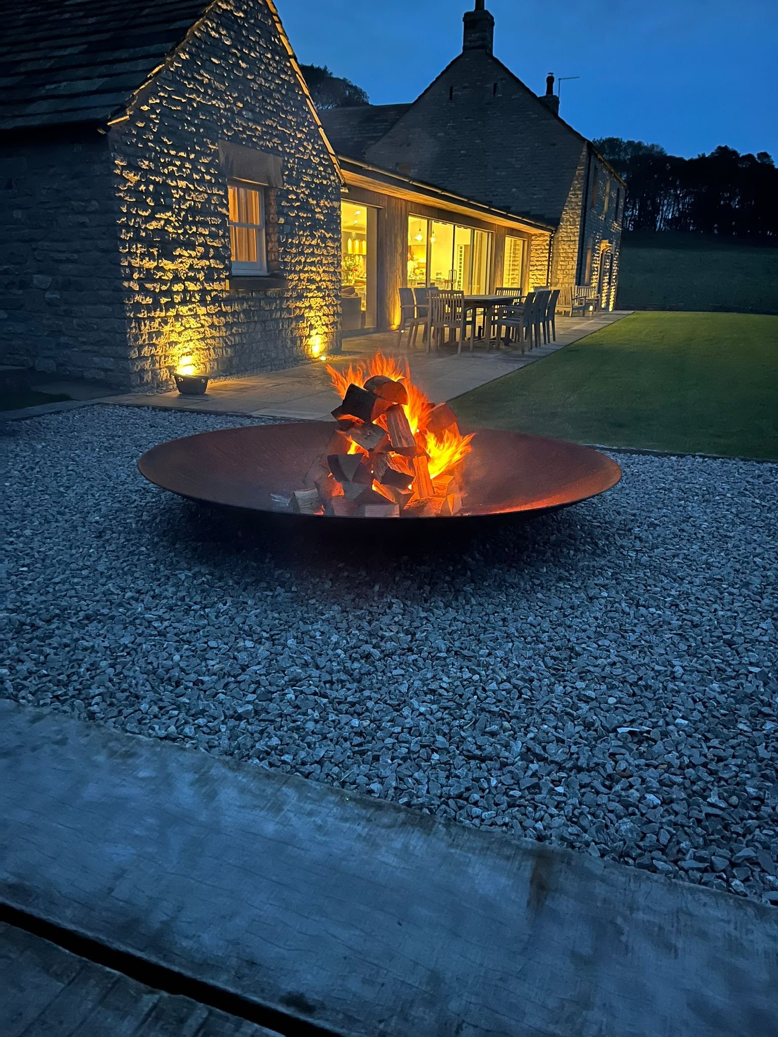 Bleaklow retreat location illuminated at night with a fire pit in the foreground on a gravel surface, outdoor table and chairs, stone exterior, large windows, and a green lawn.