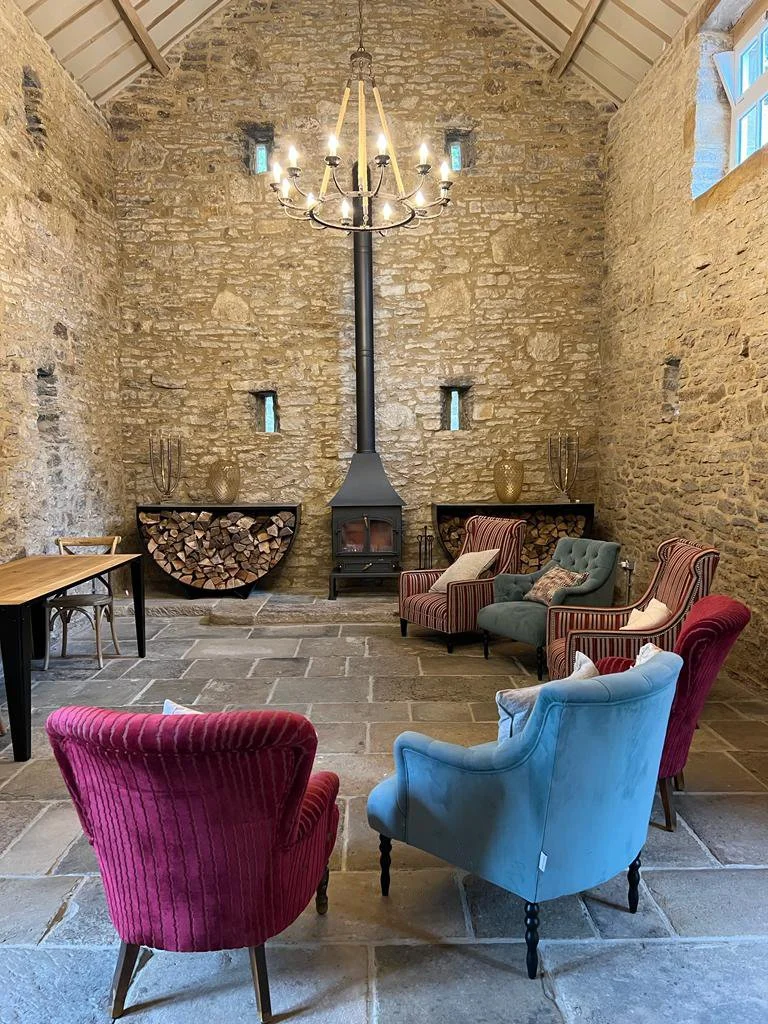 A rustic room with stone walls at our retreat space in Bleaklow, a high vaulted ceiling, a chandelier, a wood-burning stove with a pipe, a fireplace shelf with firewood, and chairs in red, blue, and green around a central point.