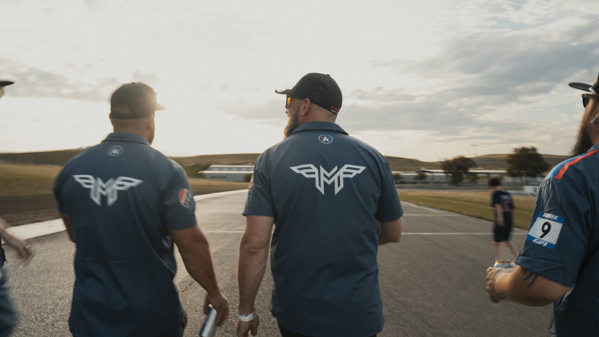 Three men wearing matching blue shirts with a logo on the back, standing outdoors on a paved area near a racetrack, with a cloudy sky in the background.