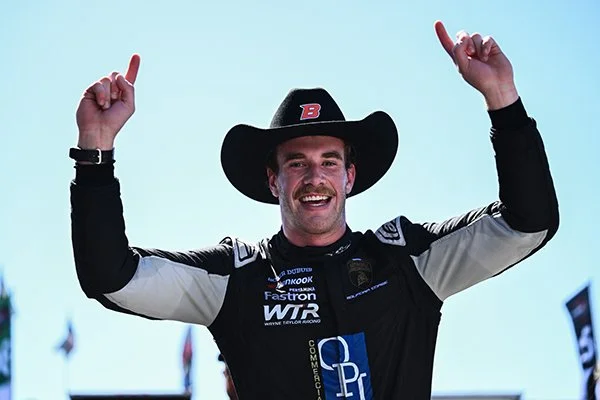 Motorsports driver celebrating victory, wearing racing suit and cowboy hat, raising arms with index fingers pointing upward, smiling. Nick Persing Wayne Taylor Racing WTR