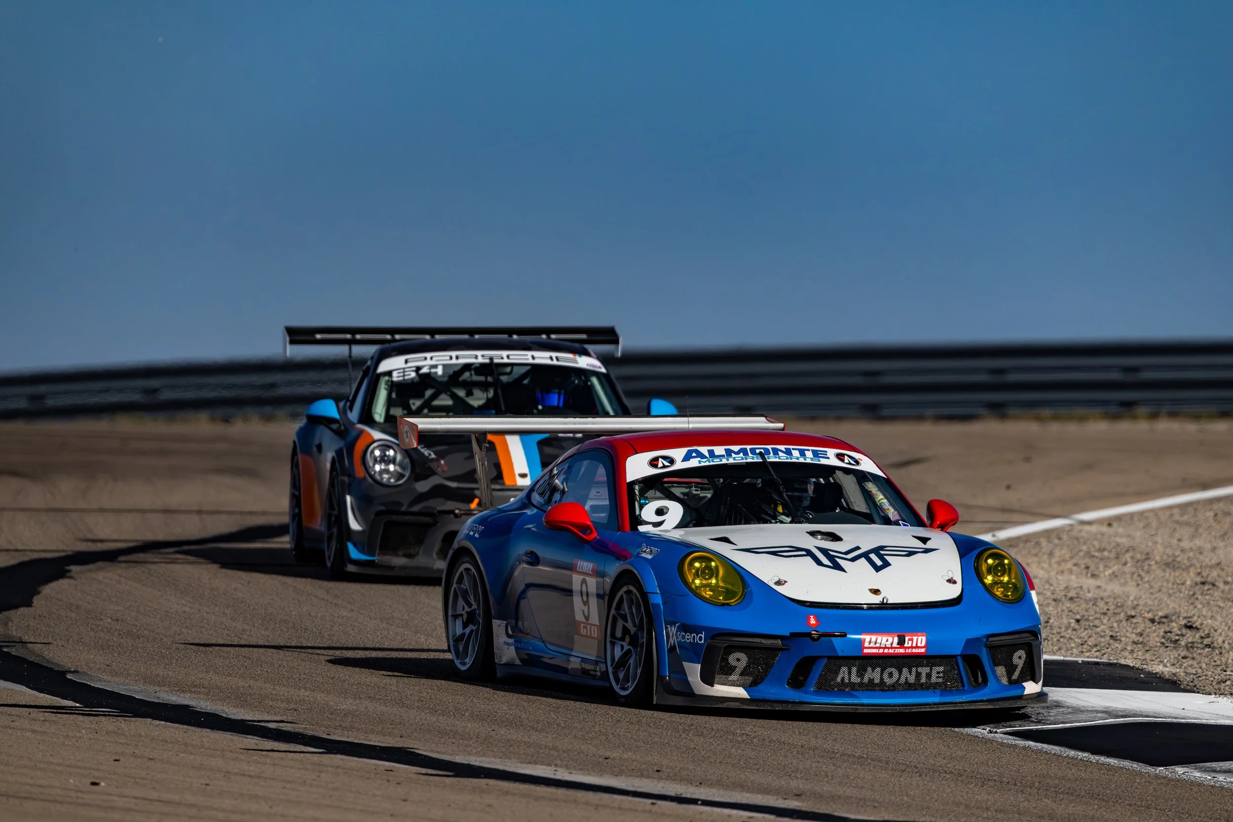 Two Porsche race cars on a track, with one blue and white car in front, followed by a gray and orange car, racing on a sunny day in Utah during the World Racing League race at West Desert.