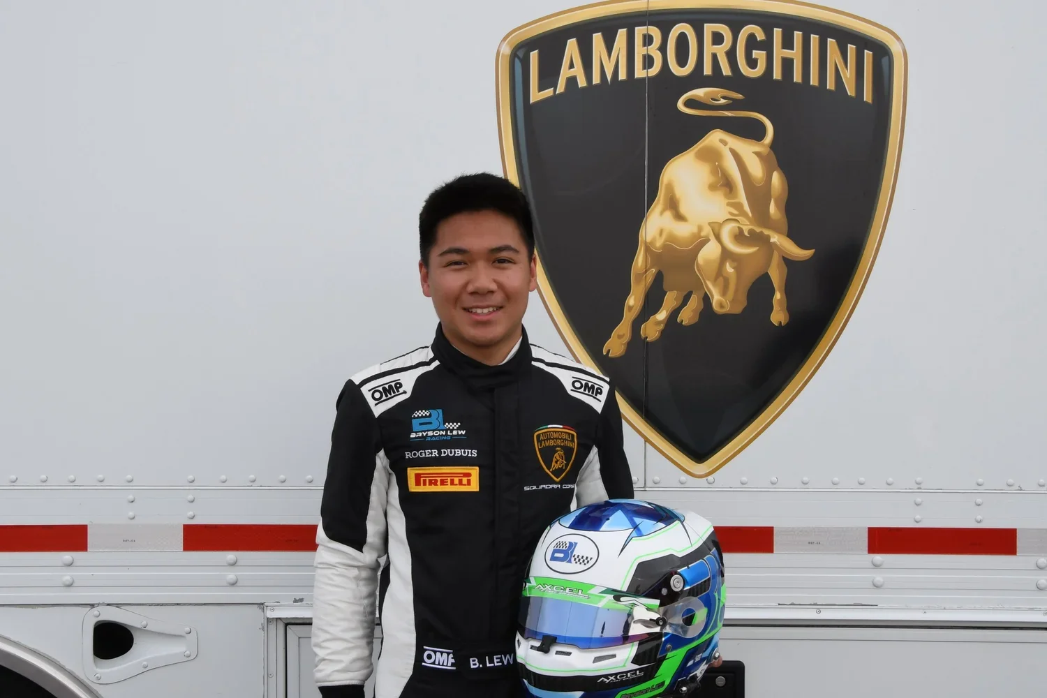 A race car driver in a black and white racing suit holding a blue, white, and green helmet, standing in front of a large Lamborghini logo on a trailer. Bryson Lew Lamborghini Super Troefo Professional