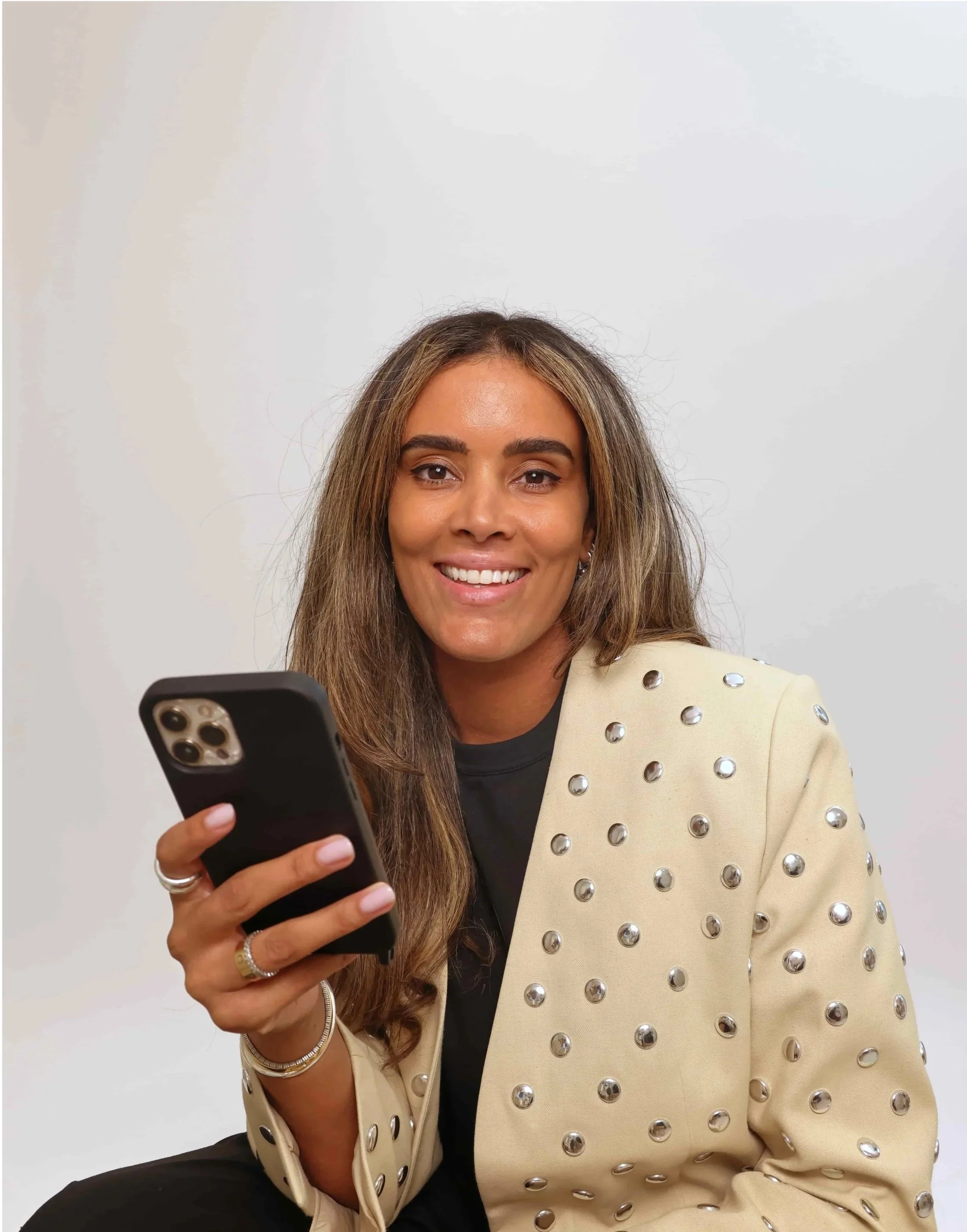 Kasey smiling with light brown hair is holding a smartphone and wearing a cream-colored jacket with metallic studs on the shoulders.