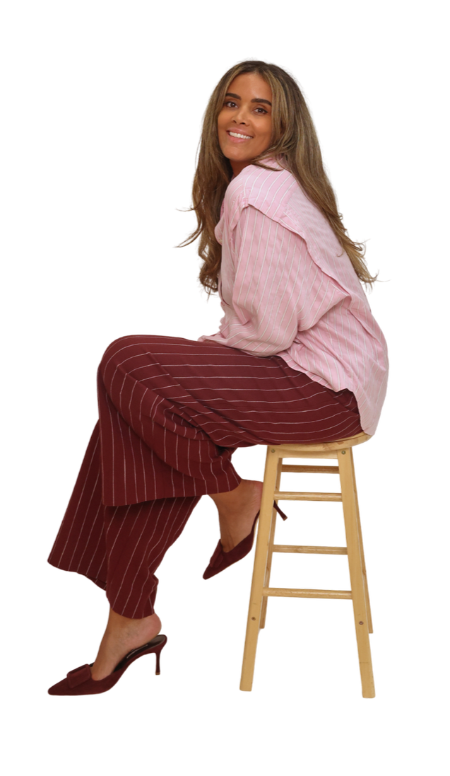A woman sitting on a wooden stool, smiling at the camera, wearing a pink striped shirt, maroon striped pants, and maroon high heels.
