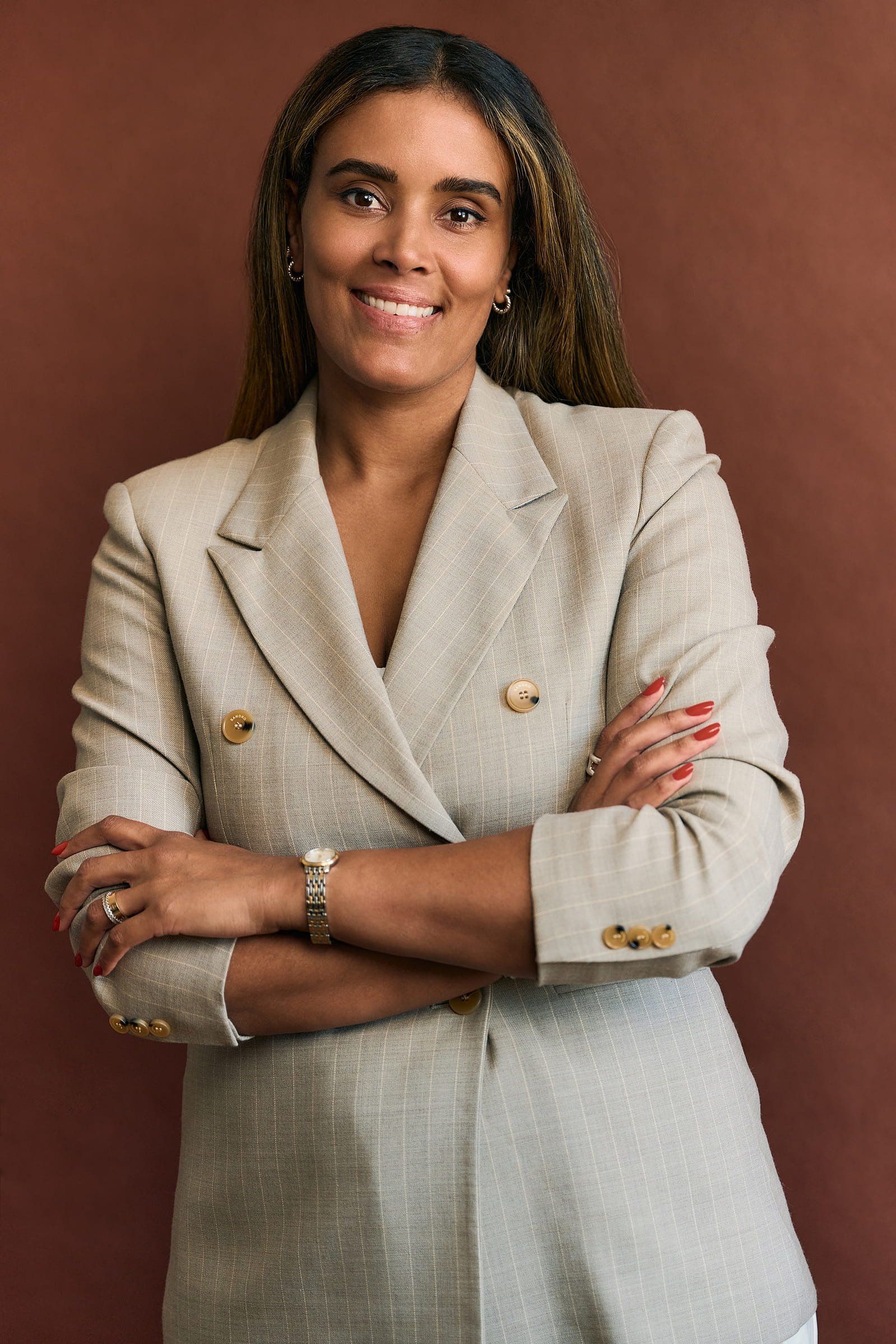 A smiling woman with long brown hair, wearing a beige plaid blazer with gold buttons and jewelry, standing with arms crossed against a brown background.