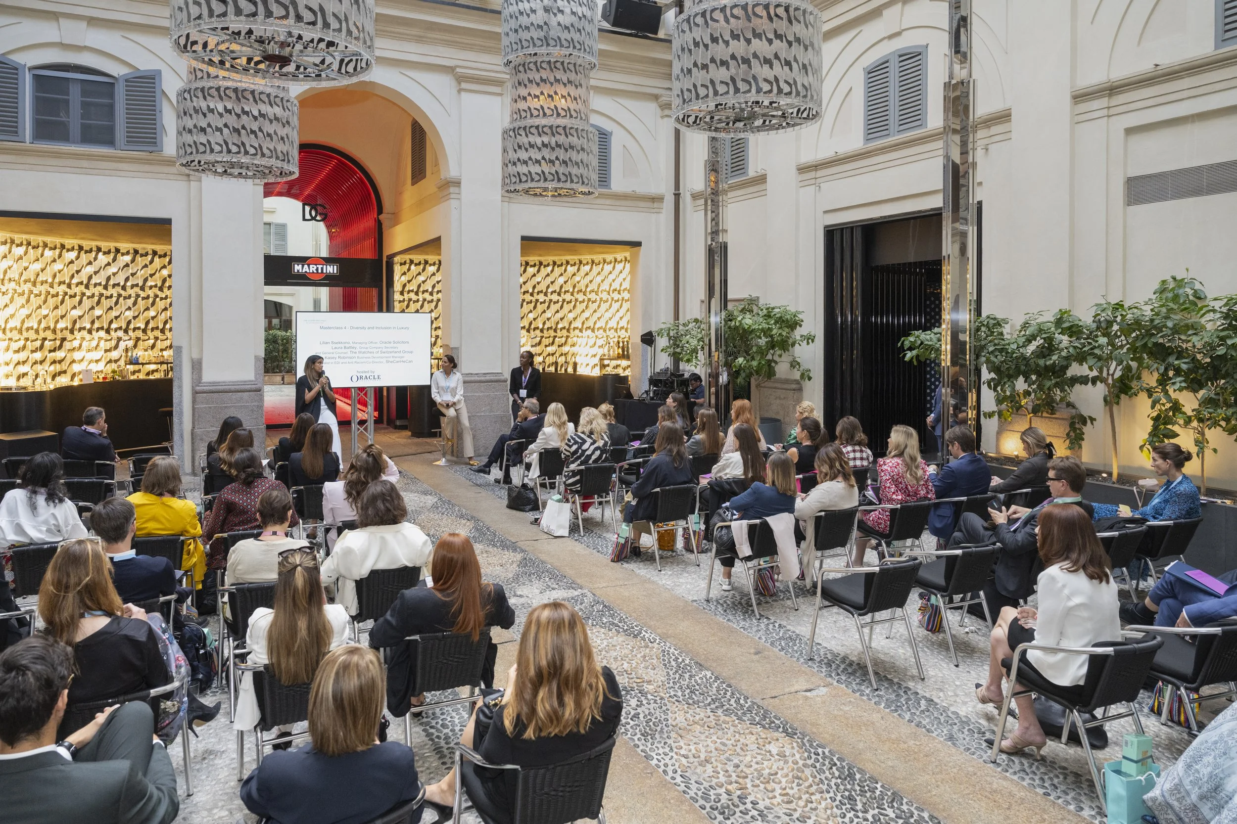 A large indoor conference taking place in a spacious, elegant venue with high ceilings and large windows. Attendees are seated in rows, facing a stage where a speaker is presenting. The audience appears engaged, and some are taking notes or looking at their devices. There is a large screen displaying presentation content. The decor includes modern lighting fixtures, greenery, and a mix of classic and contemporary design elements.