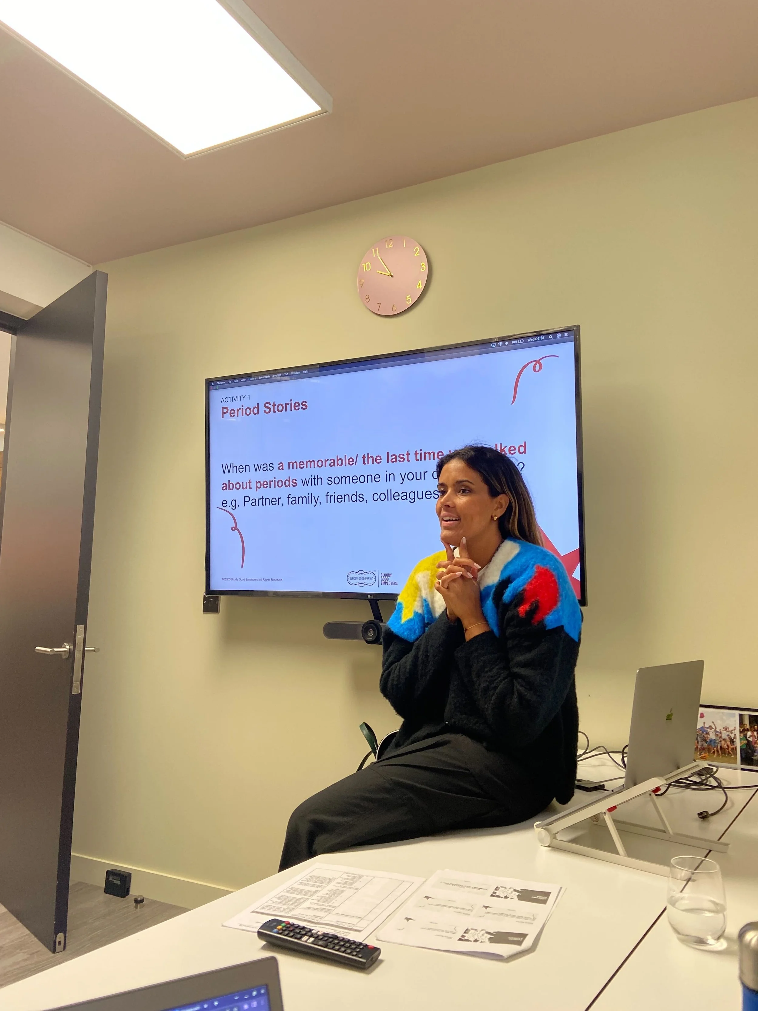 A woman sitting on a table in a classroom or conference room, leading a presentation about period stories, with a large screen behind her displaying presentation slides. There is a laptop, a glass of water, some papers, a TV remote, and a photo on the table.