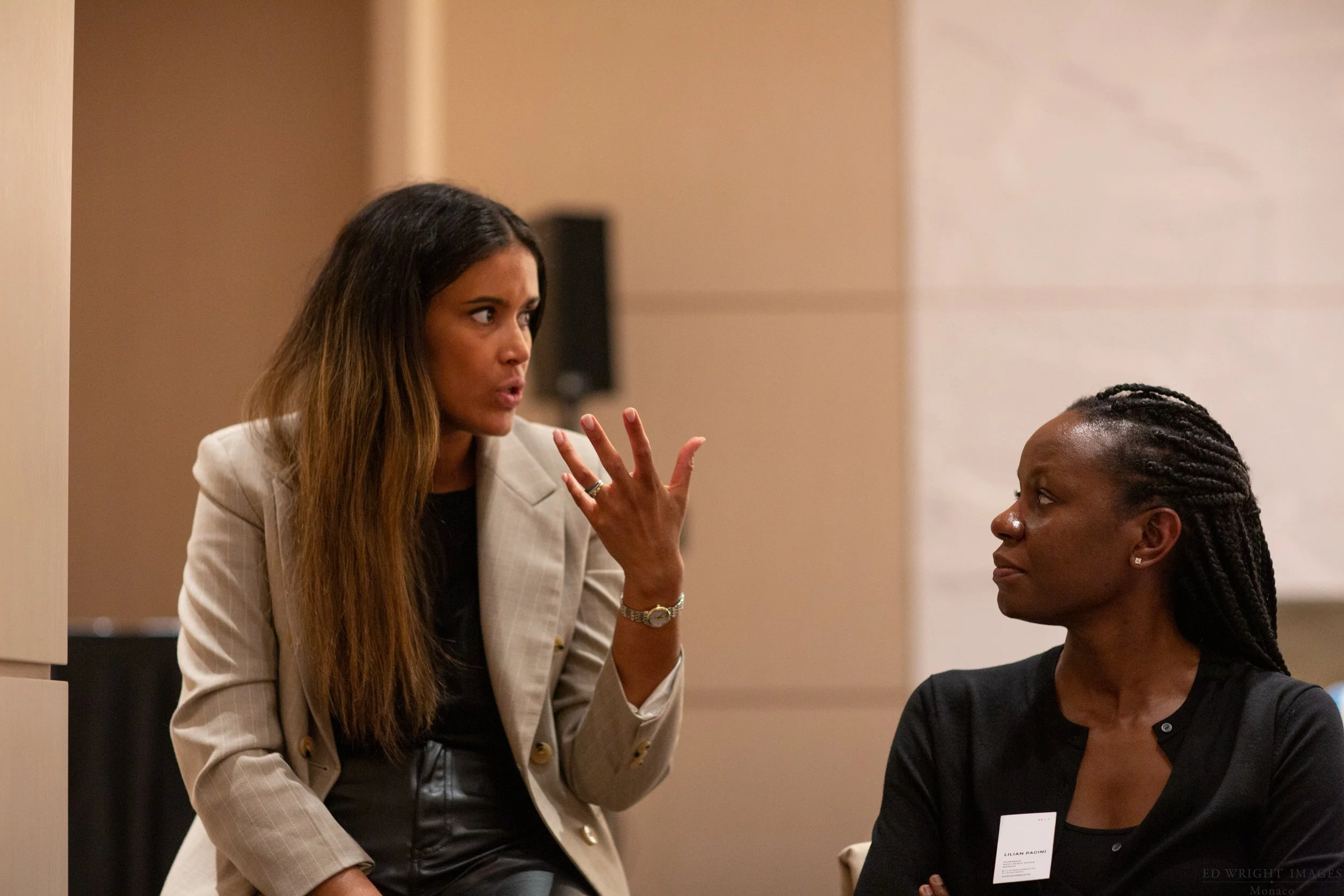 Two women engaged in a conversation: Kasey with long brown hair, wearing a beige blazer, gesturing with her hand; the other woman with braided hair, wearing a black top, listening attentively.