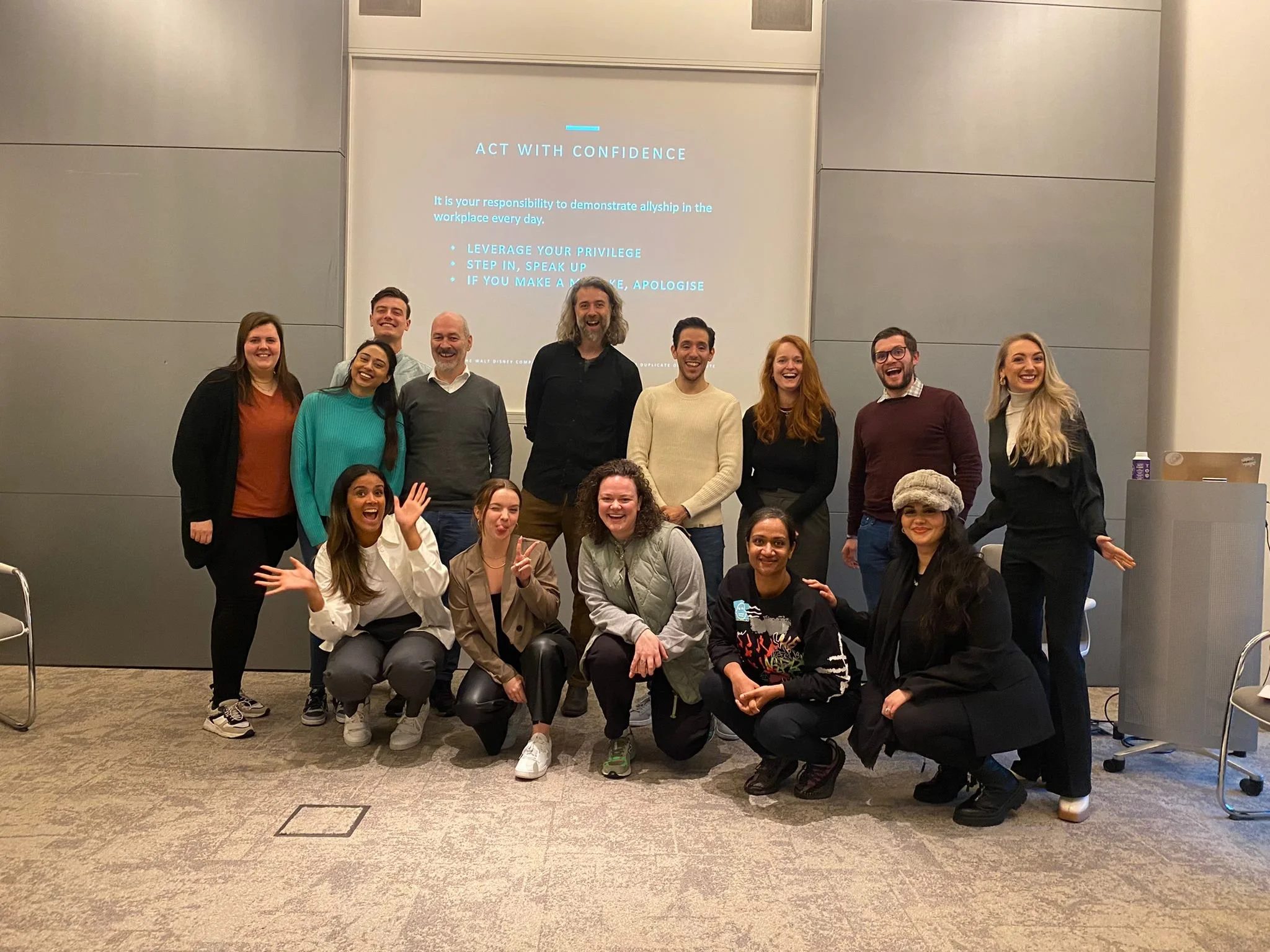 A group of 15 diverse people smiling and posing together in front of a presentation screen in a conference room.