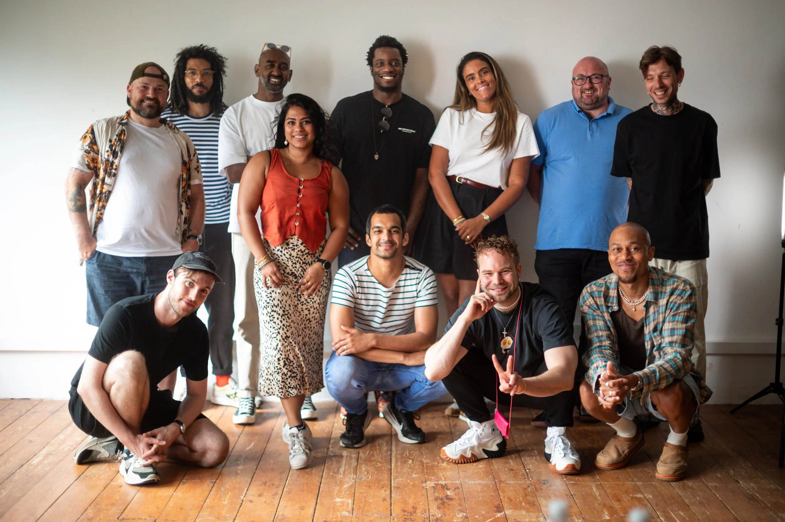 Group of 13 diverse young adults posing together indoors on a wooden floor, smiling, in casual clothing.