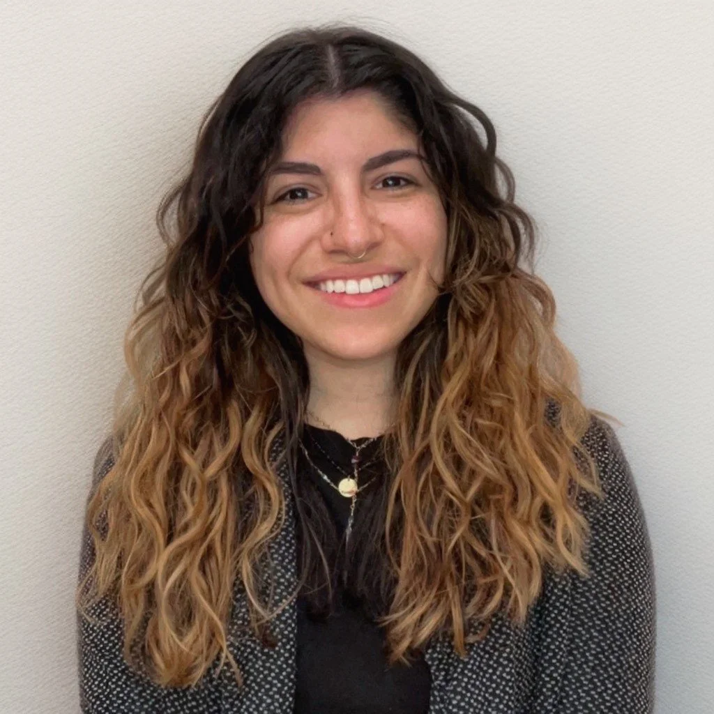 Young woman with long, curly brown hair, smiling, wearing a dark top and a gray jacket, standing against a plain wall.