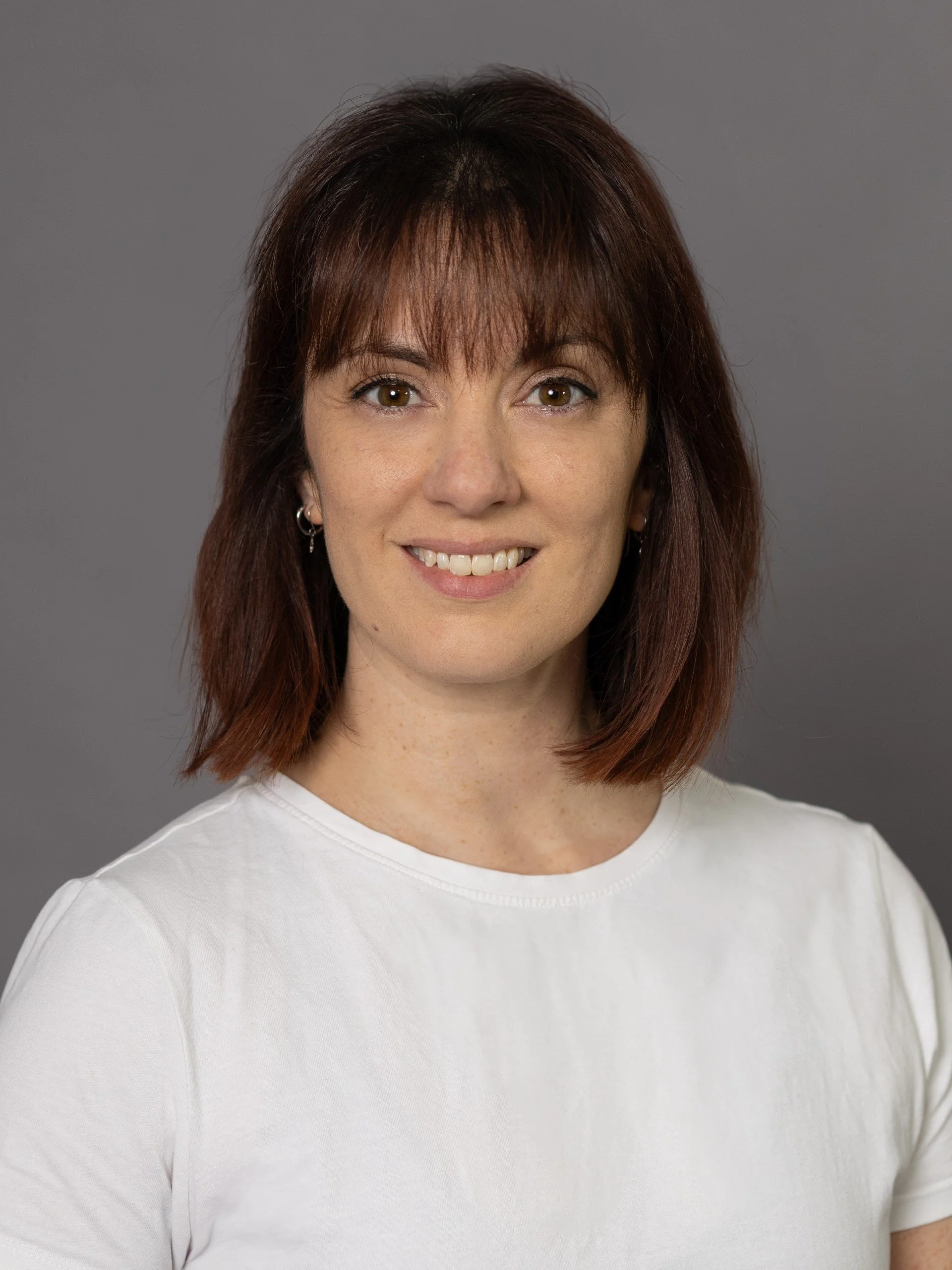 A woman with shoulder-length auburn hair and light skin smiling, wearing a white t-shirt and small hoop earrings against a gray background.