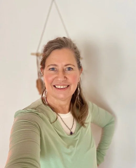 A smiling woman with light brown hair wearing a light green top and a necklace, standing indoors against a plain white wall with a decorative hanging item in the background.