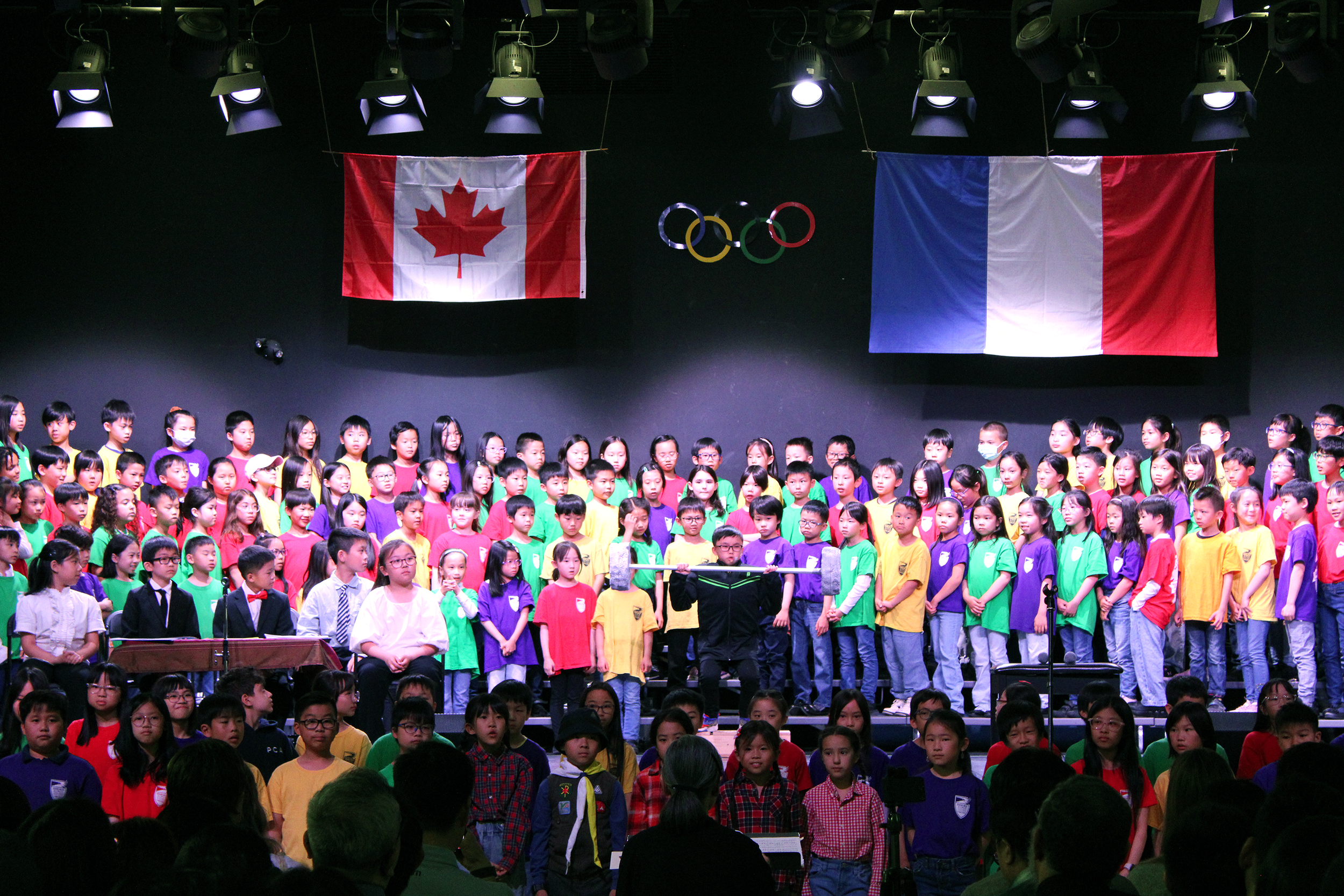 Students standing in line on stage wearing different coloured house league shirts, watching the lead actor attempt to lift the bar-bell while the council members sit at the table.