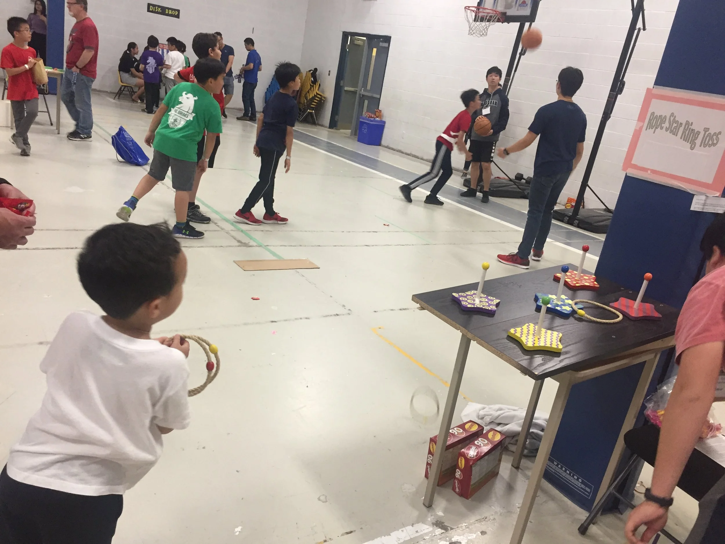 Children playing a basketball game at an indoor event, with a sign that reads 'Rope Star Ring Toss' nearby.